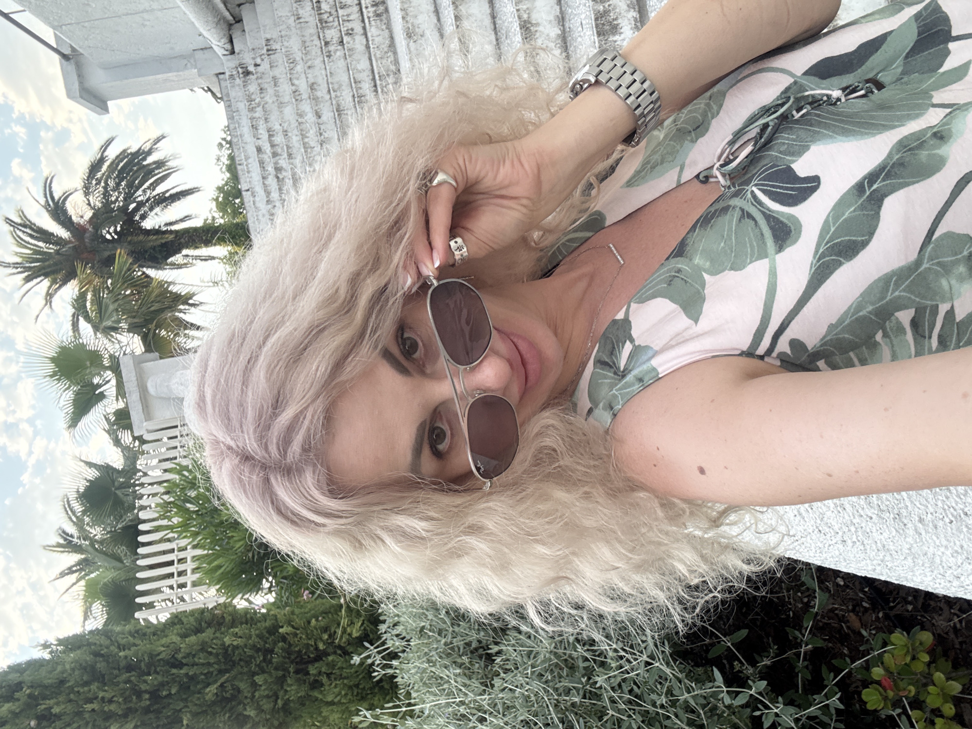 A medium shot selfie shows a woman with blonde, curly hair and sunglasses. She is wearing a sleeveless dress with a green leaf print and a silver watch on her left wrist. The woman is outdoors, with lush green foliage and palm trees visible in the background. A white fence and a stone staircase are also present, suggesting a park or resort setting. The woman is looking at the camera and holding one lens of her sunglasses. The scene is set in Sochi, Russia.