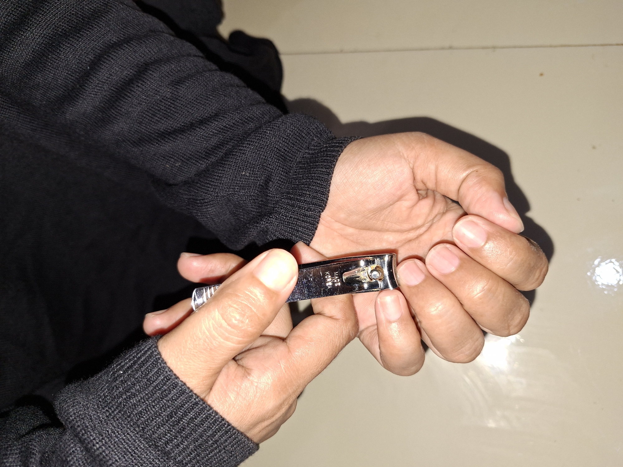 The image depicts a person, dressed in a black long-sleeved garment, using a metal nail clipper to trim their fingernails. The nail clipper is silver. The background is a light beige surface.
I can not confirm that the photo was taken in Kutaglumpang, Indonesia.