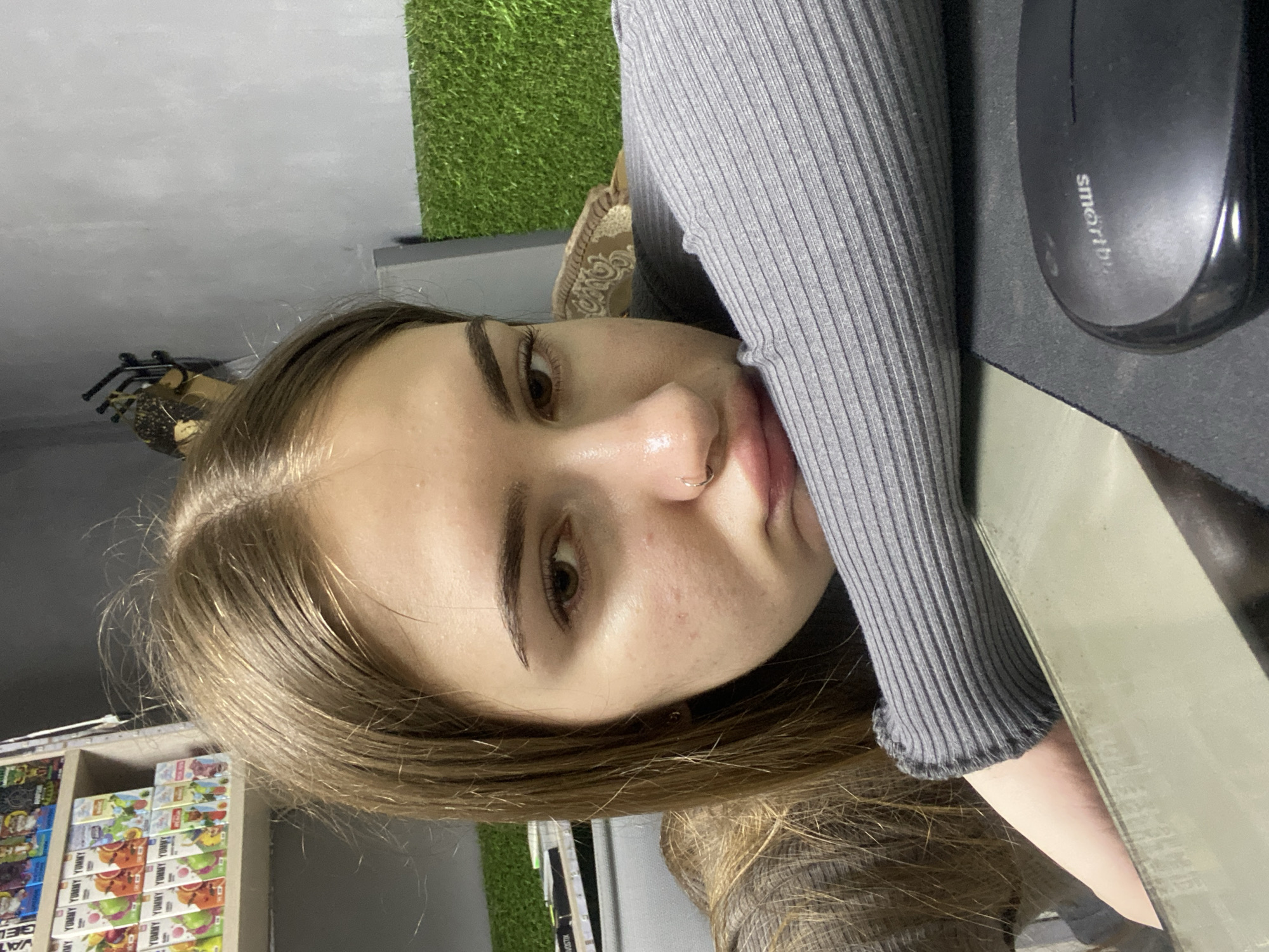 A young woman with long, light brown hair and a grey sweater is resting her head on her arm, her face turned towards the camera with a pout. She has a nose ring and her skin shows some blemishes. In the background, shelves filled with colorful juice boxes are visible, along with a patch of artificial grass. A computer mouse is also present in the foreground. The overall environment appears to be indoors, possibly a shop or a room with shelving and decorative elements. The location is noted as Dyakovo, Russia, though no specific urban features are discernible in the image.