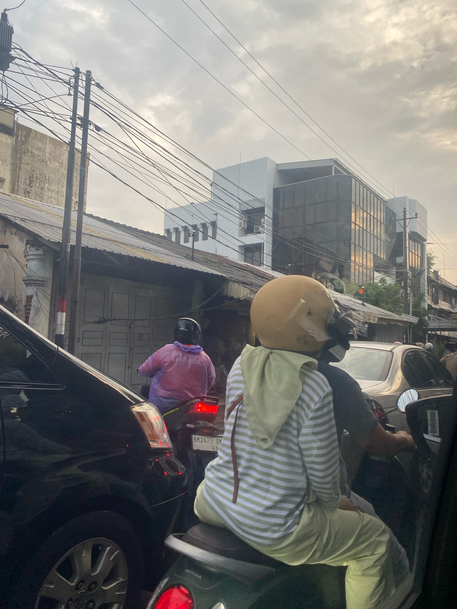 The image depicts a street scene with several vehicles and buildings under an overcast sky. In the foreground, two people are riding a motorcycle. The person in the back, wearing a yellow helmet and a green and white striped hooded shirt, is partially obscured. The person in the front, also wearing a helmet, is wearing a translucent pink poncho. The motorcycle has a red taillight and a license plate that reads "BK 2473 AN".
To the left of the motorcycle, a black car is visible, with its front fender and wheel in the lower left corner of the frame. To the right of the motorcycle, another vehicle, possibly a silver car, is partially visible.
In the background, there are several buildings. A two-story structure with a corrugated metal roof is situated behind the motorcycle. Further back, a modern, multi-story building with glass facades stands out. Electrical wires crisscross the sky, supported by utility poles. The overall impression is of a moderately busy urban environment, likely during daylight hours but with cloudy weather.