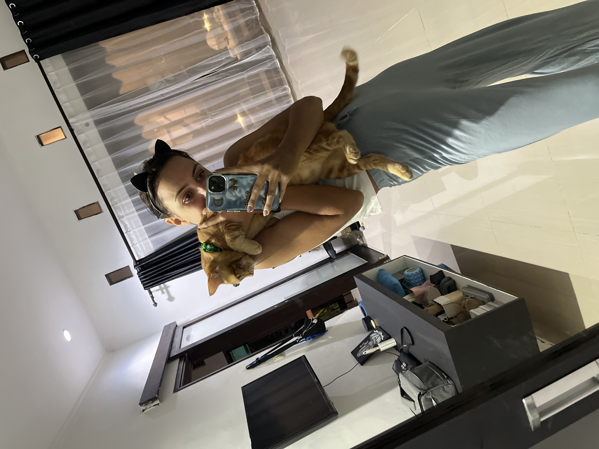 A selfie taken in Kuta Selatan, Indonesia, depicts a woman with dark hair wearing cat ears and holding an orange tabby cat. She is wearing grey pants. The woman is facing a mirror and taking a picture with her phone. Behind her are a window with a white curtain and a black curtain rod. To her right is a black desk with a computer monitor, a black box with items in it, and a grey backpack. The floor is a light beige color.