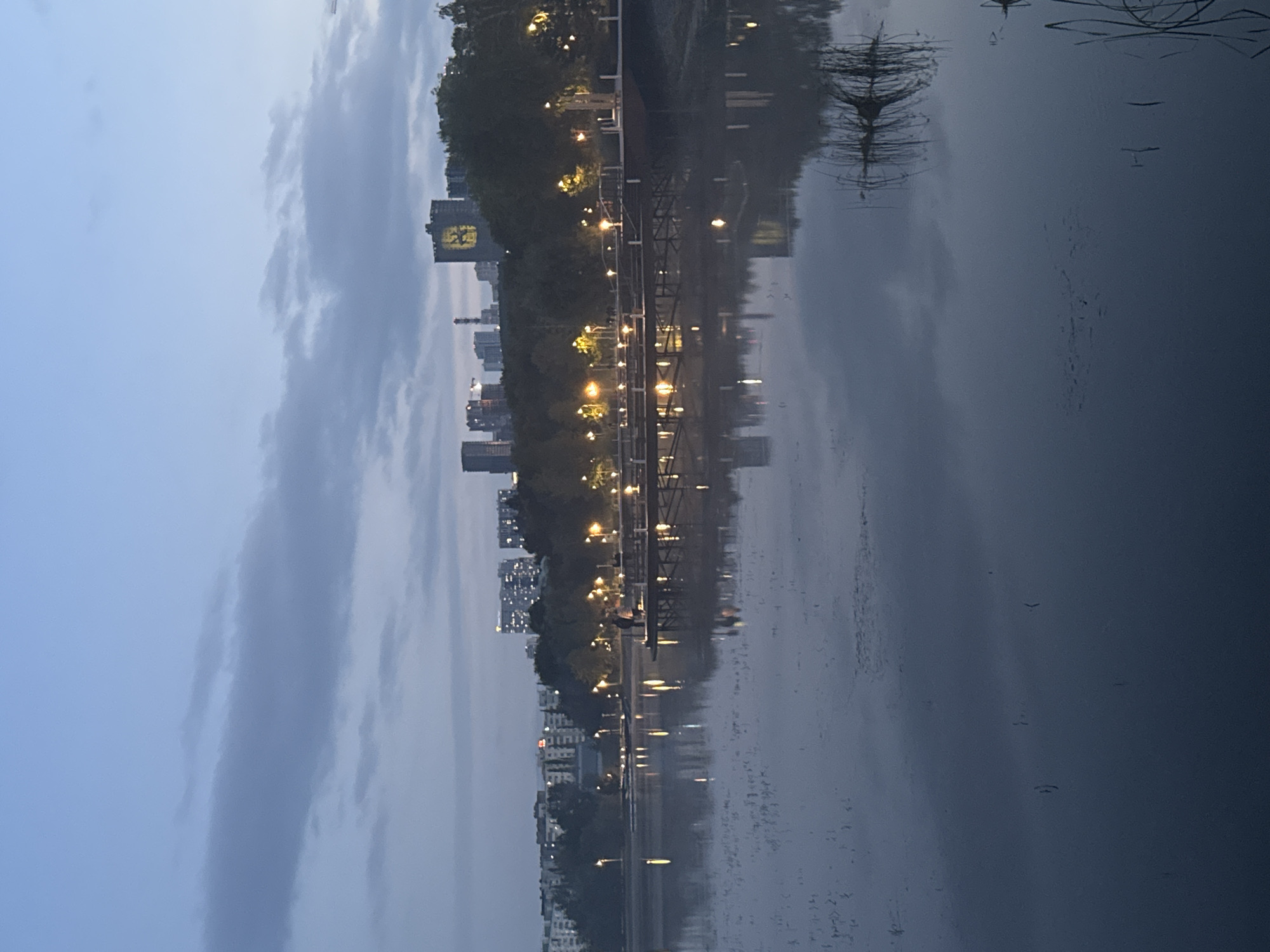 The image depicts an evening scene at the Ufa Arena, specifically the area surrounding the Ufa River embankment, as indicated by the Moscow State University emblem visible on one of the buildings. The sky is a deep twilight blue, with scattered grey clouds. A calm body of water, reflecting the dim lights of the city, occupies the foreground and middle ground.
In the middle distance, a wooden pier extends into the water. Several lampposts line the pier and the embankment, casting a warm yellow glow. Two figures are visible on the pier, appearing to stand and interact. Beyond the pier and embankment, a dense line of trees forms a dark silhouette against the cityscape.
The background features a collection of multi-story buildings, characteristic of an urban environment. Prominently displayed on one of the taller buildings is a large emblem, resembling the Moscow State University logo, illuminated in yellow. The overall atmosphere is tranquil and serene, typical of an urban park at dusk.