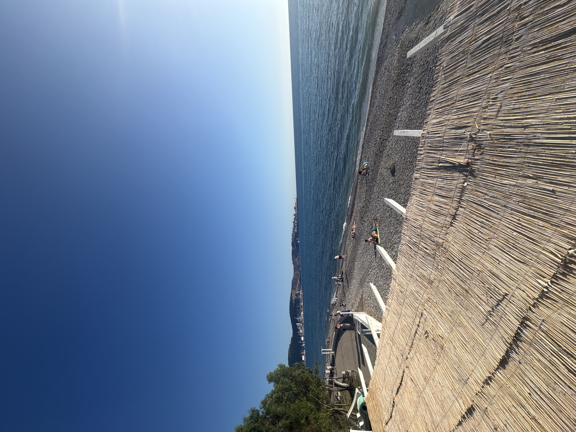 The image depicts a panoramic coastal scene in the city of Bar, Montenegro, under a clear, bright blue sky with a slight solar glare in the upper right. In the immediate foreground, a textured, light-brown thatched or woven bamboo-like canopy or roof extends across the lower portion of the frame, supported by visible white concrete or painted structural elements.
Below these structures, a pebble beach, predominantly grey and brown, stretches along the coastline. Approximately 15-20 individuals are scattered across the beach. Some are lying prone on towels or mats, presumably sunbathing, while others are standing or walking. For example, one person in green attire is lying on a mat near the bottom right.
To the left, green trees with dense foliage partially obscure a paved, dark-grey promenade that runs parallel to the beach. A light blue-green pedal boat or similar small recreational watercraft is partially visible on the left side of the promenade.
The deep blue sea occupies the midground and extends to the horizon, exhibiting small, visible waves near the shore. Across the bay, in the background, the distinct skyline of a city is visible at the base of undulating hills or mountains. This city features numerous buildings and, notably, a prominent port facility with multiple large cranes and several ships docked, identifying it as the port area of Bar, Montenegro.