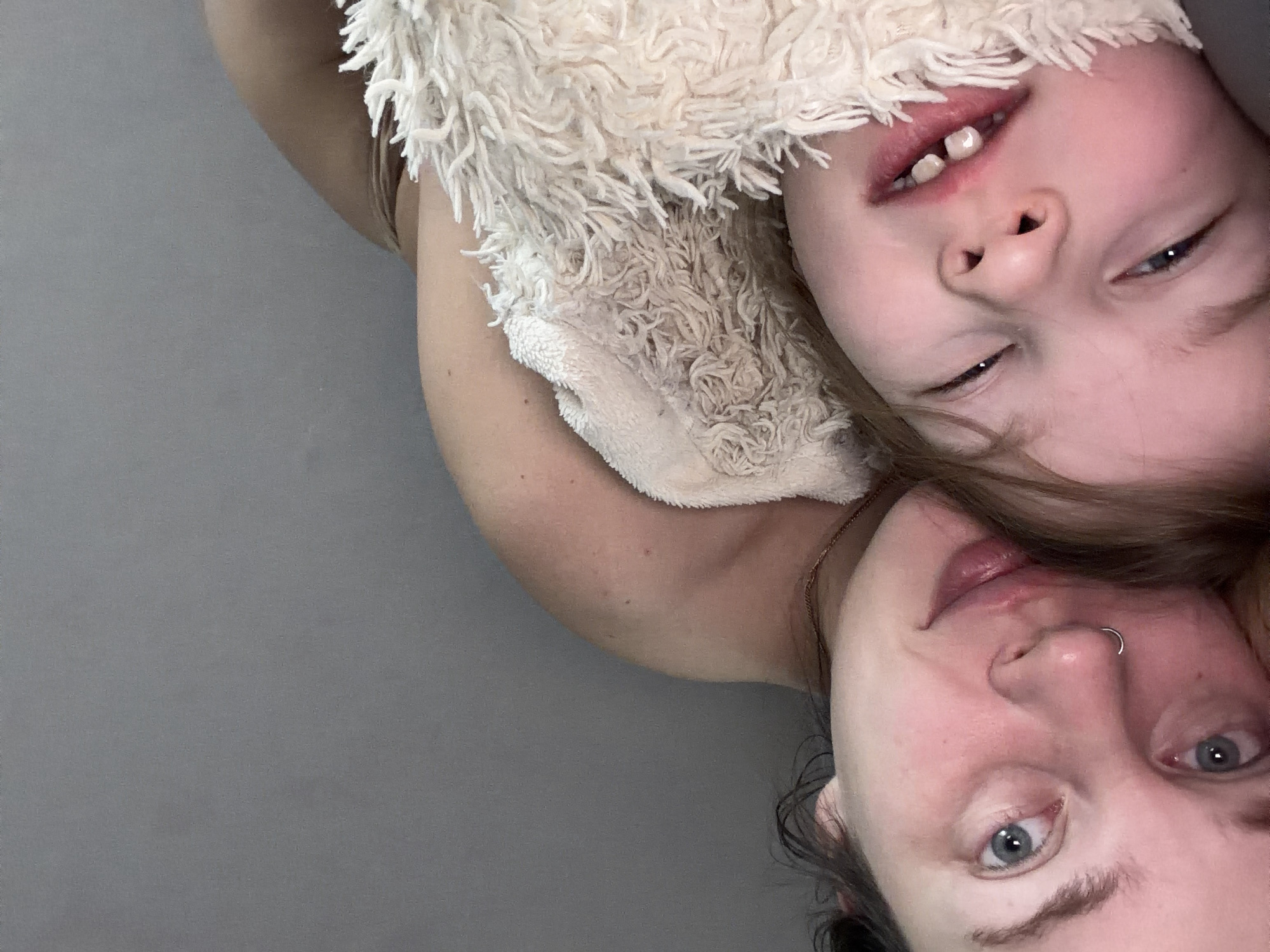 A woman and a young child are lying down together, facing the camera, and angled at 180 degrees. The child, who appears to be a toddler, is partially covered with a soft, fluffy, white material, possibly a blanket or garment. The woman has a nose ring and blue eyes. They are lying on a plain, gray surface. There is no indication of the location being Chaikovsky, Russia, or any urban setting.