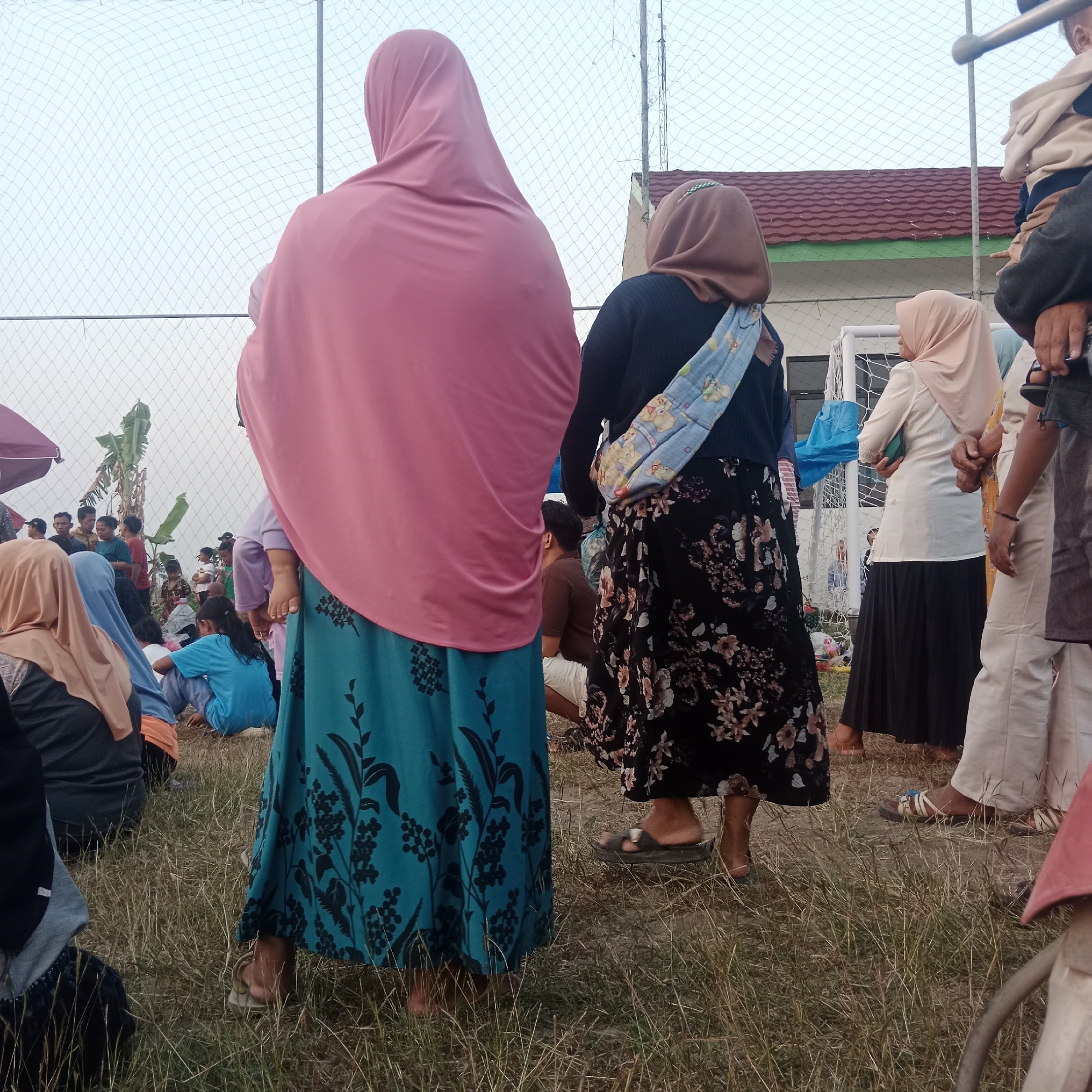 In Tegowanu Wetan, Indonesia, a group of people are gathered outdoors, possibly for an event or a casual social occasion. The setting appears to be a grassy field adjacent to a fenced area with a goalpost, suggesting a sports field or recreational space. The sky is overcast, and the time of day seems to be late afternoon or early evening.
In the foreground, two women are the primary subjects, viewed from behind. The woman on the left wears a vibrant pink hijab and a teal skirt with a black floral pattern. She is holding a baby, visible from the back, whose head is resting on her shoulder and is partially covered by the hijab. The woman on the right wears a brown hijab and a black skirt adorned with a detailed floral print in shades of pink and white. She has a colorful baby sling draped across her body.
Behind them, other individuals are visible. A child in a light-colored hooded garment is being held by an adult. Further in the background, more people are scattered across the field, some sitting and others standing. A small building with a red tile roof and green trim is partially visible behind the fencing. Banana trees are also present in the background, adding to the tropical atmosphere. The overall scene depicts a communal gathering in an outdoor environment.