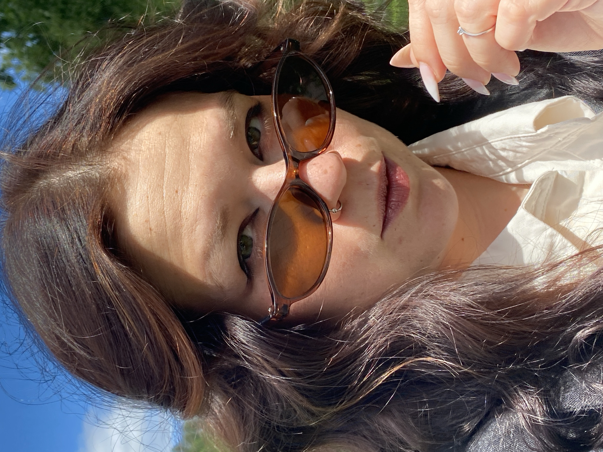 The image shows a close-up, outdoor portrait of a woman with long, dark, wavy hair. She is wearing brown-tinted, oval-shaped sunglasses and a light-colored collared shirt. A silver ring with a small stone is visible on her right ring finger. She has a nose ring on her left nostril. The background is blurred, showing green foliage and a blue sky with some clouds. The lighting suggests it is daytime. There are no indications that this image is located in the city of Chaikovsky, Russia.
