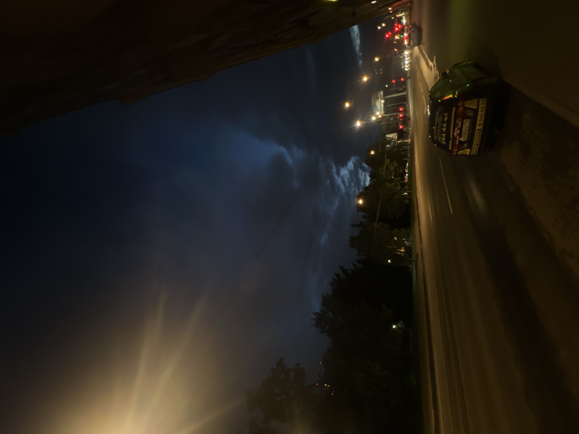 A nighttime street view of Saint Petersburg, Russia, captures a dark, cloudy sky with dramatic light rays emanating from an unseen source on the left. The street is illuminated by streetlights and the headlights of passing cars, creating a reflective wet surface. On the right side of the road, a dark building looms, its texture indistinct due to the low light. A yellow taxi with Cyrillic lettering and a phone number "344 44 44" is visible on the right lane of the street, moving away from the camera. Trees line the left side of the street, silhouetted against the dimly lit sky. Traffic lights can be seen in the distance, indicating an intersection ahead.