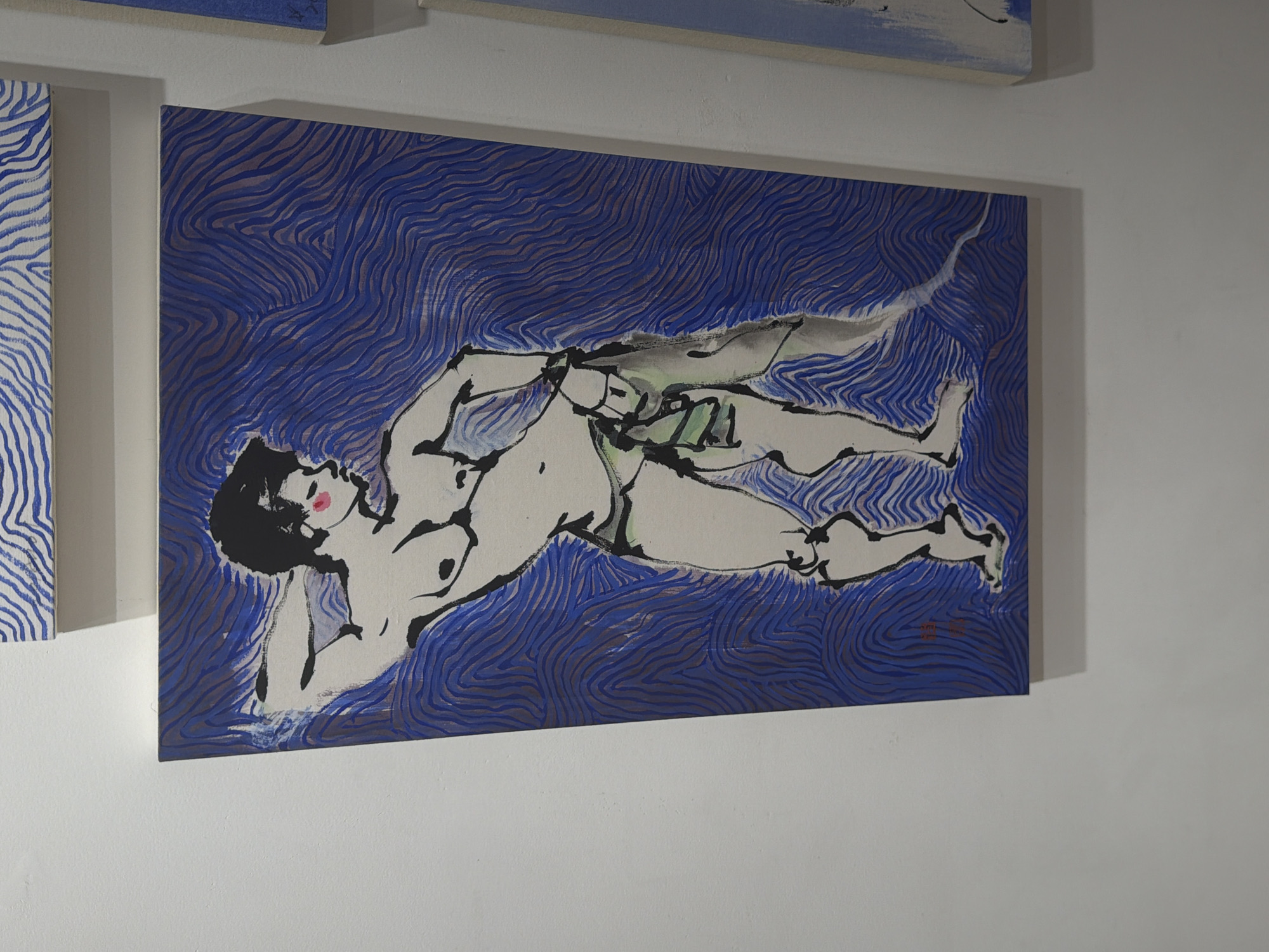 The image shows a painting of a male figure in a dynamic pose, rendered in an expressive, gestural style with bold black outlines and flat areas of color. The background is a vibrant blue with swirling, wave-like patterns. The figure appears to be wearing a green cloth around its waist and has blush marks on its cheeks. The painting is displayed on a white wall, and part of another painting is visible to the right, which has a similar blue background. The scene is likely in an art gallery or exhibition space. Based on the context of its exhibition, this artwork is located in Seoul, South Korea.