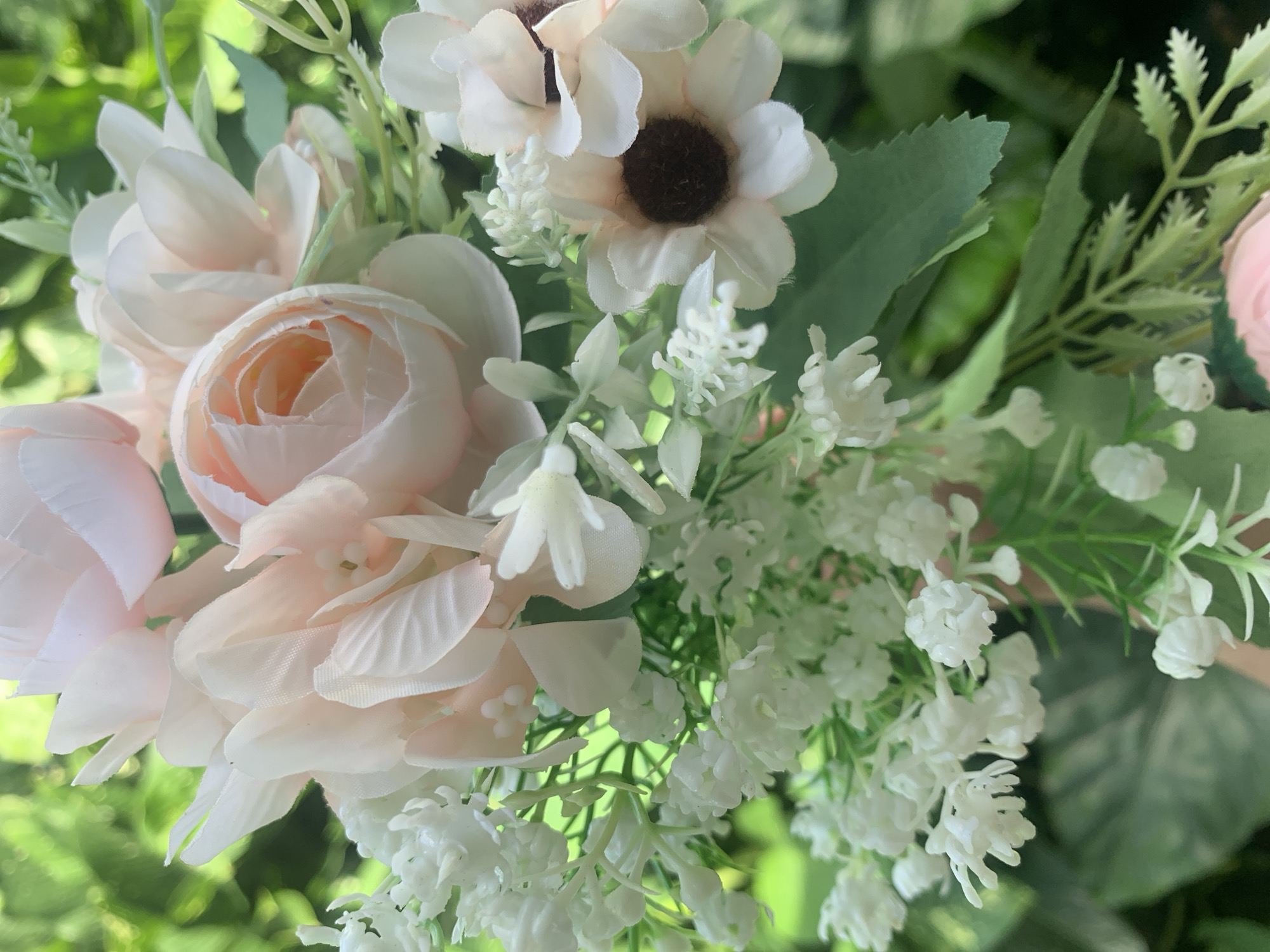 A close-up, vertical, medium shot captures a bouquet of artificial flowers outdoors. The bouquet consists of pale pink roses with layered petals, interspersed with small clusters of white, baby's breath-like flowers and feathery green foliage. One pale pink flower with a dark brown center, resembling a daisy, is visible on the right side. The background is a soft, out-of-focus blur of lush green leaves, suggesting an outdoor setting, possibly a garden or park. The image is well-lit, with natural light highlighting the textures and colors of the flowers. The location is not specified in the provided image, and thus cannot be determined to be Kuala Pembuang, Indonesia.