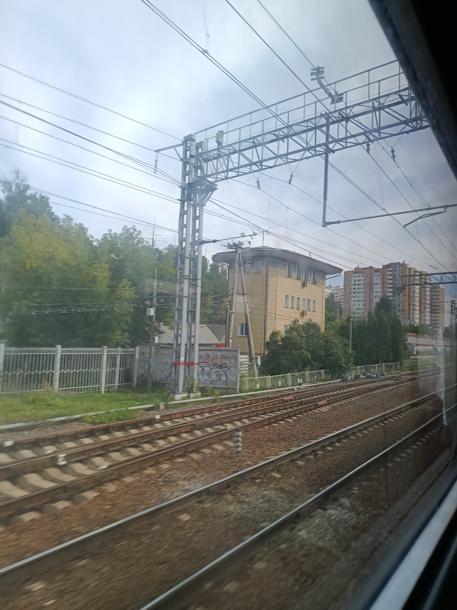 This image captures a view from a train traveling through Balashikha, Russia. The foreground is dominated by multiple sets of train tracks running parallel to each other, composed of gravel ballast and metal rails. In the mid-ground, a silver metal fence runs along the left side of the tracks, separating them from a grassy area. Beyond the fence, a light brown, two-story building with numerous windows and air conditioning units is visible. Further in the background, several multi-story apartment buildings with orange and brown facades line the horizon. The scene is framed by overhead electrical wires and supports for the railway infrastructure. The sky is overcast, contributing to a muted natural light. No individuals are clearly visible.