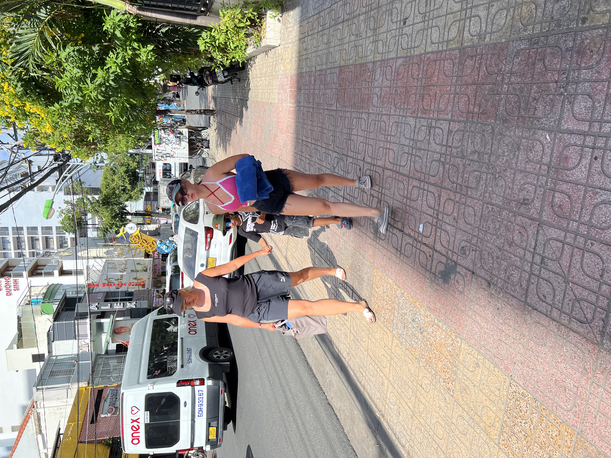 This image depicts a street scene in Nha Trang, Vietnam, likely during daytime given the bright sunlight casting shadows. The focus is on two individuals, a woman in the foreground wearing a black tank top and shorts, and another woman behind her in a pink and white halter-neck top and black shorts. Both women are wearing sunglasses, and the woman in the foreground has a baseball cap on. They appear to be standing on a patterned tiled sidewalk next to a white van.
The van, a "XE HỢP ĐỒNG" (contract vehicle), is branded with "anex" and features license plates starting with "50H-933.67". Other vehicles are visible on the street, including a white car with a license plate "79H 3.06" and various motorcycles parked in the background.
The background reveals a bustling street with buildings displaying signs for businesses like "SPA" and "ALEX". Lush green trees with yellow flowers are present, adding a touch of nature to the urban environment. Electrical wires are strung across the scene, common in Vietnamese cityscapes. The overall impression is a snapshot of daily life on a sunny day in a Vietnamese city.