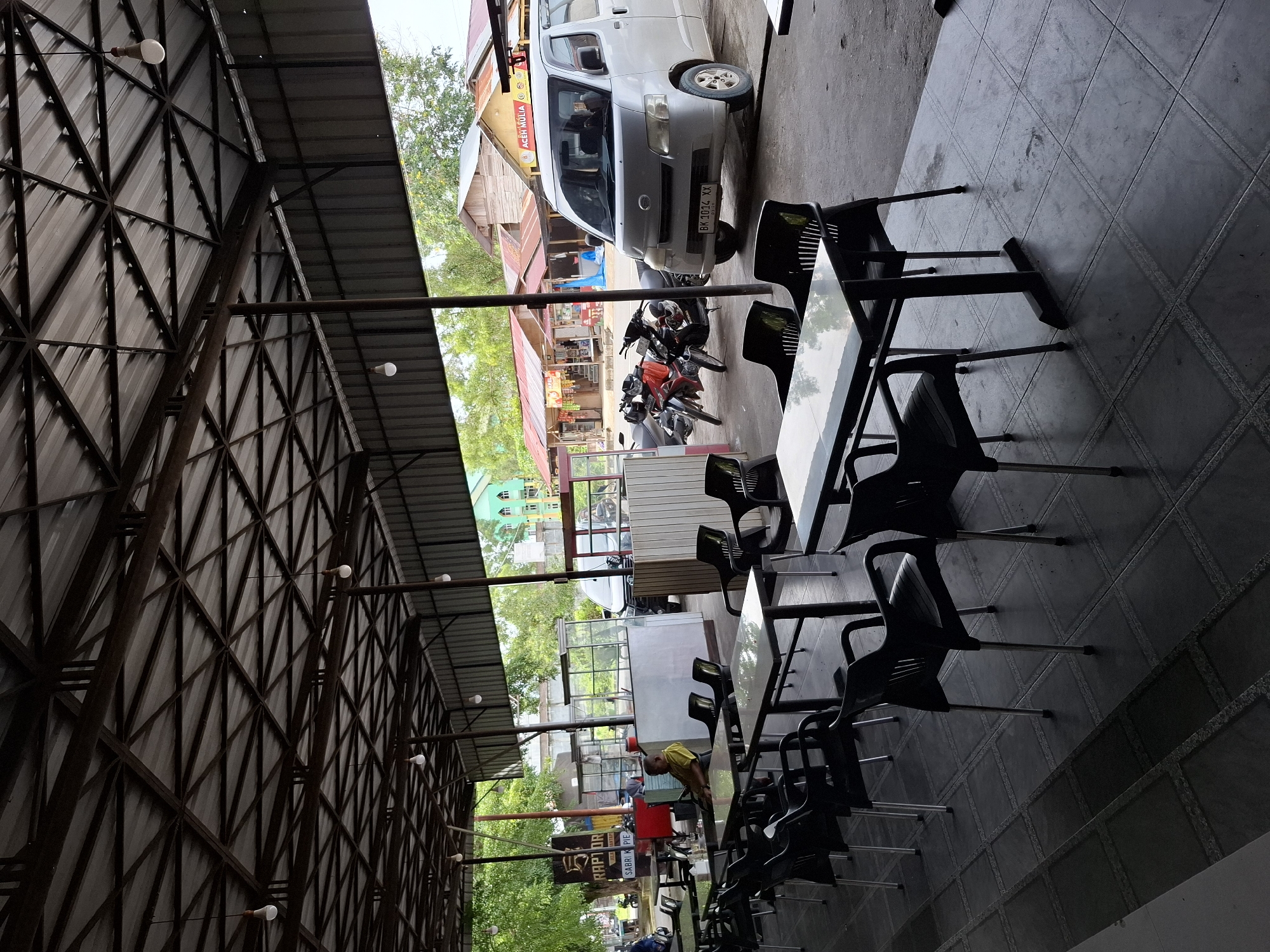The image depicts an outdoor dining area in Lhokseumawe, Indonesia. Tables and chairs are arranged on a tiled floor. Overhead, a metal roof structure is visible, with bare light bulbs hanging from it. A silver van is parked outside the dining area, along with two motorcycles. Across the street, buildings and trees can be seen. A person wearing a yellow shirt is present within the dining area. A sign with "RAPTOR" is visible.
