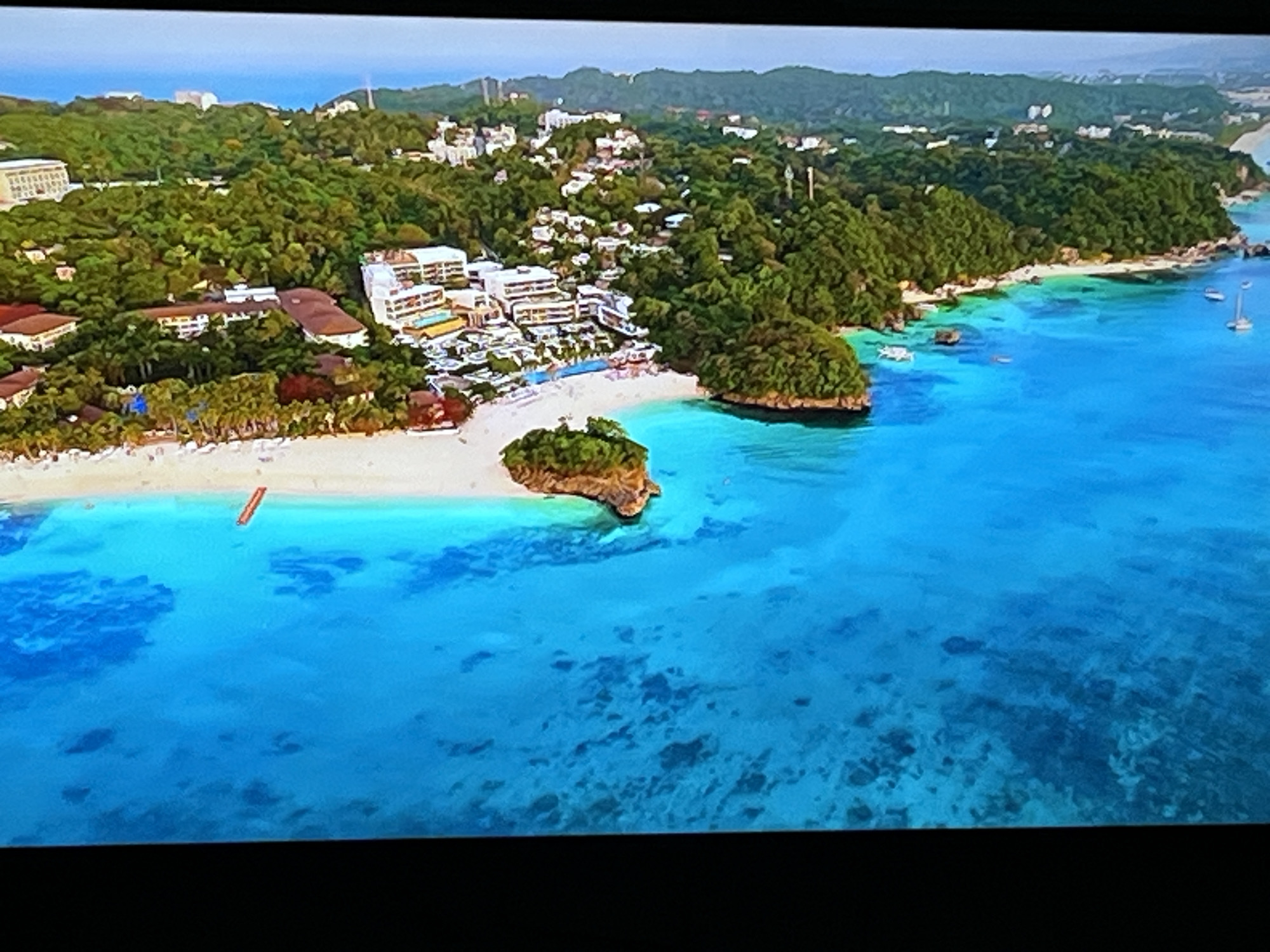 This is an aerial, oblique photograph of a tropical island resort. The foreground is dominated by clear, turquoise water with visible coral formations beneath the surface. A small, rocky island with lush green vegetation is situated in the mid-ground, close to a white sandy beach. The beach is lined with palm trees and several resort buildings with white and brown facades. In the background, a densely vegetated hillside rises, dotted with more buildings and houses. The sky above is a pale blue, with a hazy horizon. There are no people or animals visible in the scene. The location is not Kharkiv, Ukraine, but rather a tropical coastal area, likely a beach resort.