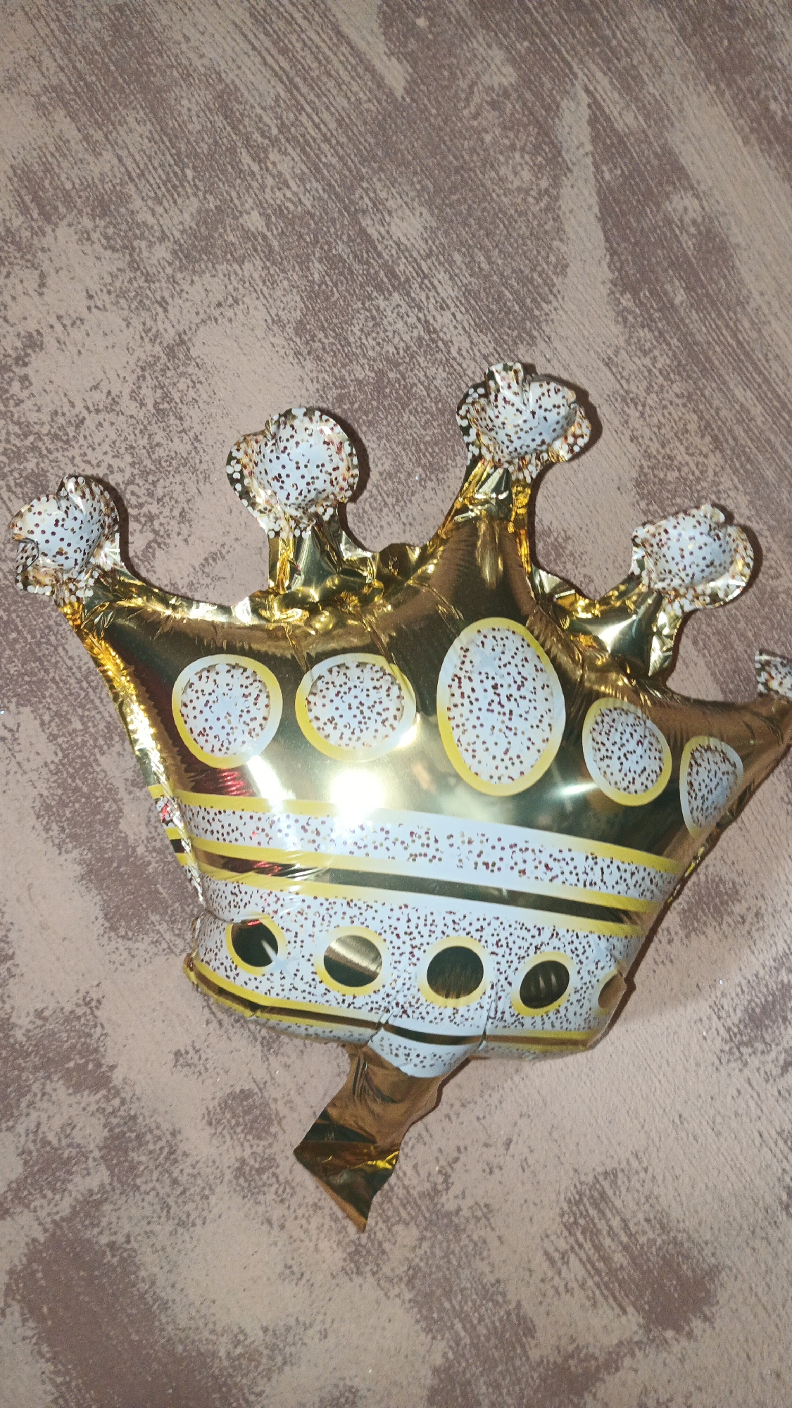 A gold foil balloon shaped like a crown is lying on a textured, taupe-colored surface, possibly a wall or fabric backdrop. The balloon is decorated with white bands containing gold circular accents filled with small, reddish-brown dots. The crown has five points, each tipped with a white, dotted accent. The background surface has a distressed, brushed appearance. The scene is illuminated by a light source coming from the left, casting reflections on the shiny balloon. There are no people or other objects visible in the image. The location is not specified in the image, and there is no information available to suggest it is in Kryvyi Rih, Ukraine.