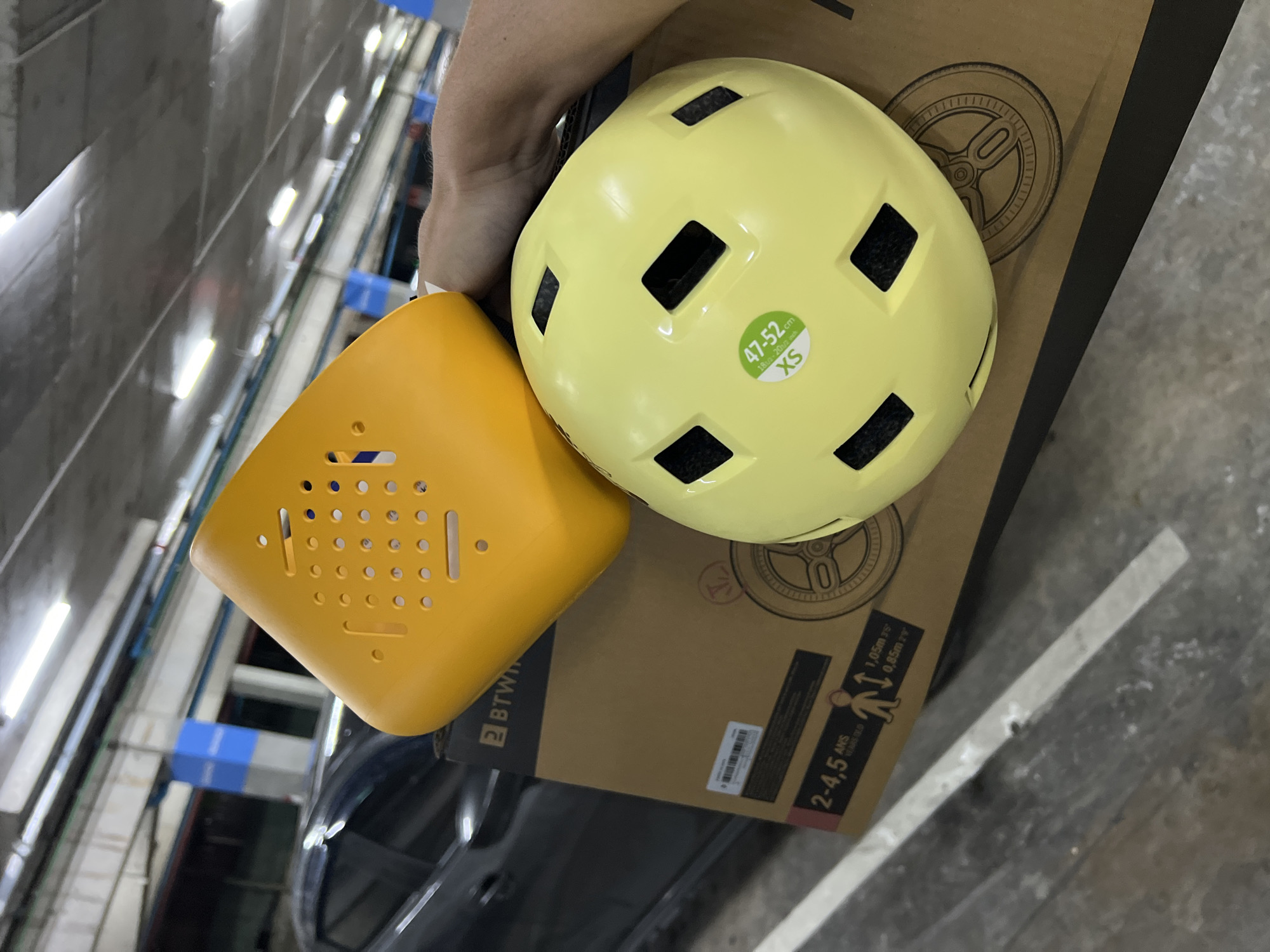 The image captures a scene from an elevated perspective, showcasing a hand holding a yellow child-sized bicycle helmet and a small orange plastic basket, likely for a bicycle. The helmet features ventilation holes and is labeled with a size specification of "47-52cm XS." These items are positioned on a cardboard box with printed graphics, including the text "2-4,5 ANS" and illustrations related to bicycle use. The background suggests an indoor or covered parking structure due to the presence of overhead lighting, metal beams, and a visible portion of a vehicle. The photograph appears to be taken in Unknown, Unknown, as the image lacks details about the specific city.