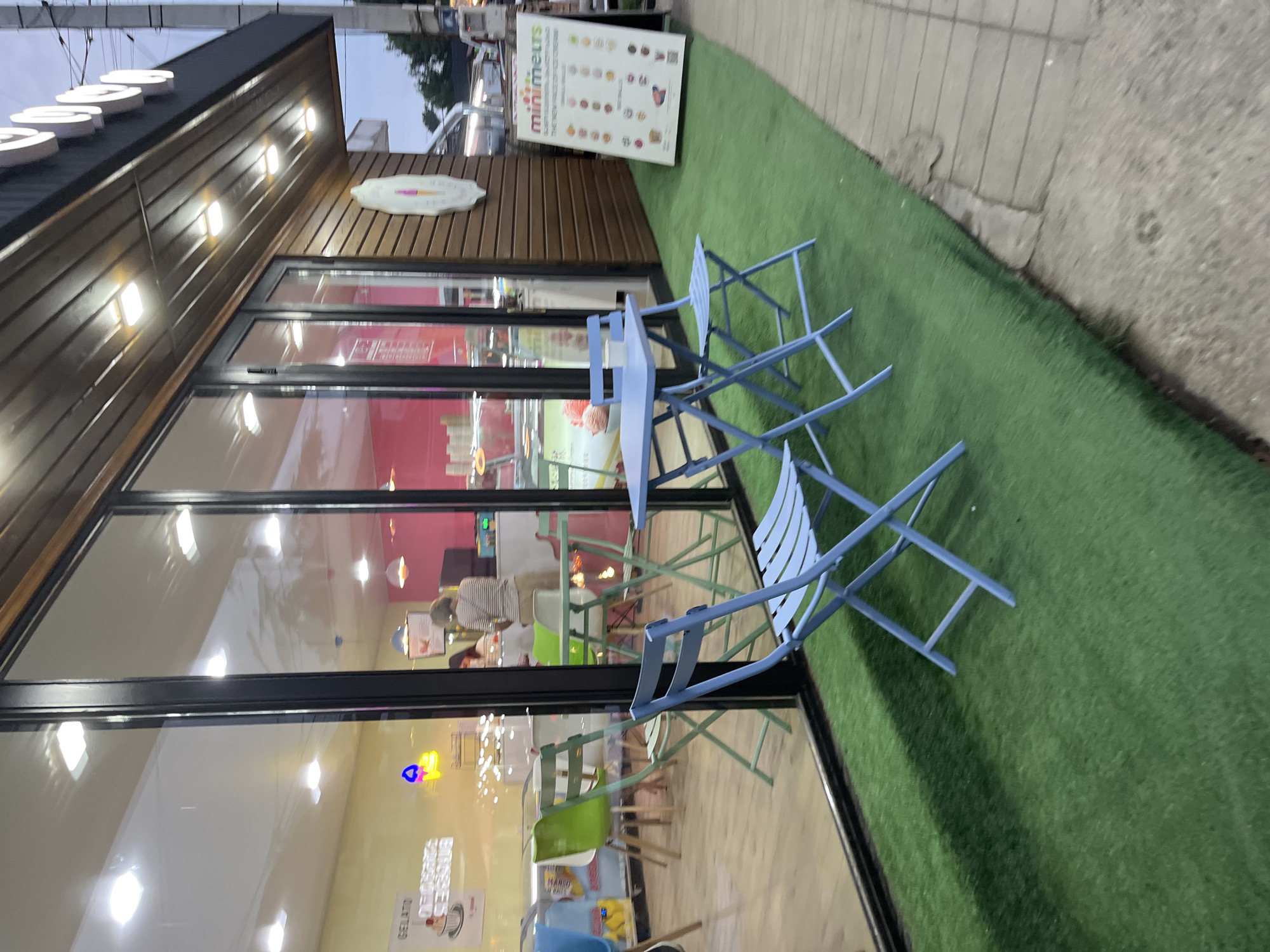 The image depicts the exterior of a "Mini Melts" ice cream shop in Ureki, Georgia. The shop has large glass windows revealing a brightly lit interior with pink walls and display cases. Outside, there are several light blue folding tables and chairs set up on artificial turf, suggesting an outdoor seating area. A sign for "Mini Melts" is visible, featuring pictures of their ice cream products. The architecture of the shop includes wooden paneling above the windows and a dark awning. In the reflection of the glass, a person can be seen inside the shop, appearing to be an employee. The overall scene suggests a pleasant, casual establishment, likely popular with tourists and locals alike in the seaside resort town of Ureki.