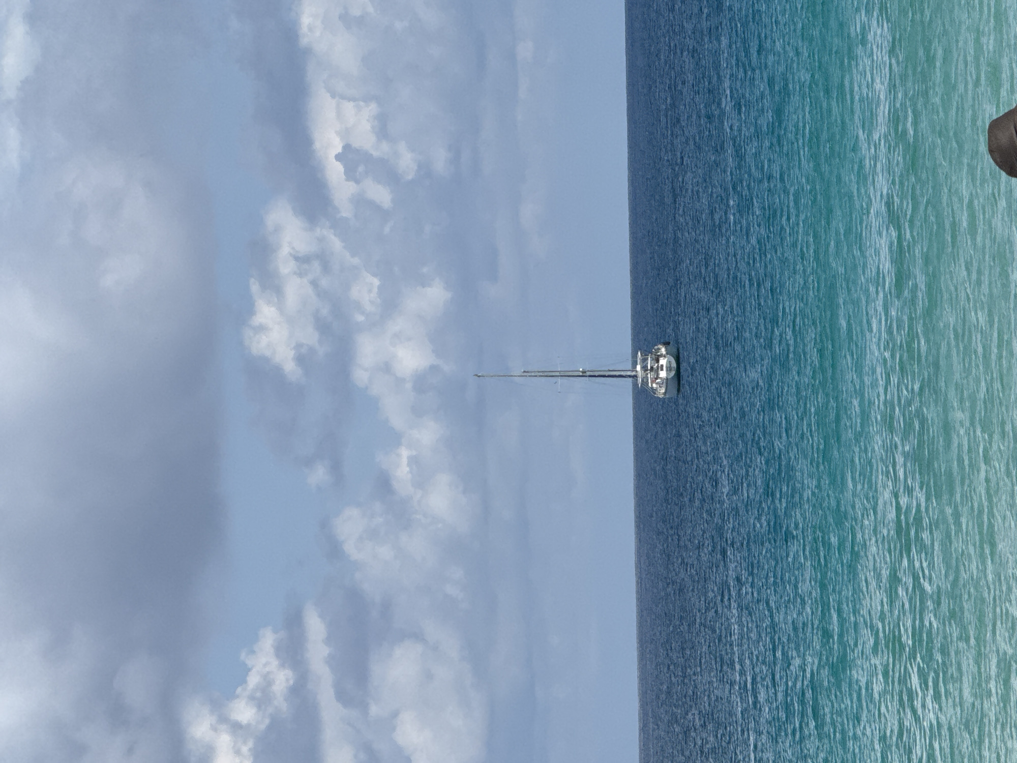An overcast sky with scattered white and gray clouds hangs over a calm, deep blue ocean. A single white sailboat with a tall mast is positioned centrally in the frame, anchored in the water. The ocean transitions from a darker blue in the distance to a lighter, turquoise hue closer to the foreground. The water is slightly rippled, indicating a gentle breeze. The scene is devoid of any landmass or other vessels.