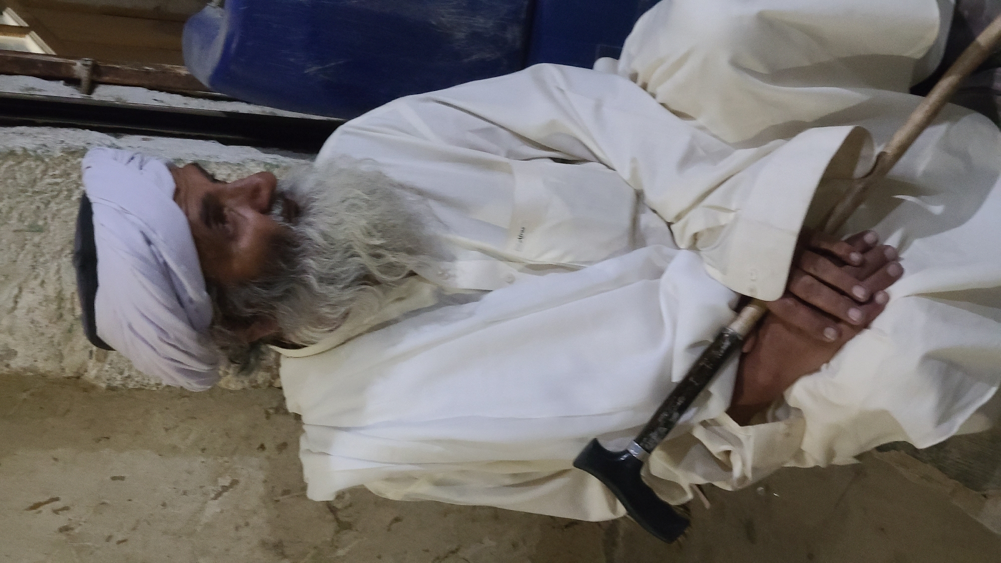 An elderly man lies on his side on a rough, pale surface. He wears a white turban and a long white robe. His long white beard is prominent. A dark walking cane rests between his folded hands. A blue container is visible in the background. The location is Ziarat, Pakistan.