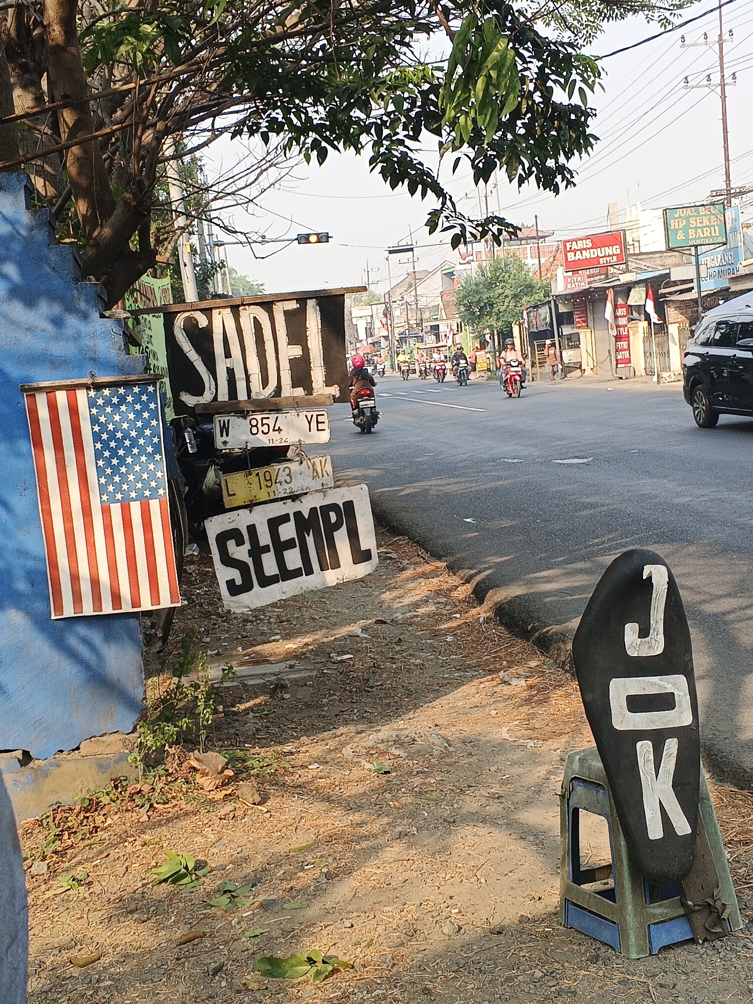 The image depicts a street scene in Tambak Kemerakan, Indonesia, on a sunny day. In the foreground, to the left, a blue wall is visible, with an American flag displayed on it. Next to the flag are several signs. The largest sign reads "SADEL" in white block letters on a black background. Below "SADEL" are two license plates. The top license plate has the characters "W 854 YE" and "11-24". The bottom license plate has "L 1943 AK" and "11-22-24". A third sign below these reads "STEMPL" in black block letters on a white background. To the right of the signs, a black motorcycle seat with the white letters "JOK" is propped up on a blue and green plastic stool.
The street itself is paved and appears to be a main road with moderate traffic. Several motorcycles are seen on the road, with riders wearing helmets. In the background, there are buildings lining the street, some with red signage that includes text such as "FARIS BANDUNG STYLE" and "FITRI BATIK". Other signs on the buildings include "JUAL BELI HP SEKEN & BARU" and "PAKARANSI HP MURAH". Traffic lights and utility poles are also visible. The overall environment is urban, with a mix of commercial establishments and residential buildings. The ground is a mix of dirt and gravel. Trees with green leaves are present in the upper part of the image, casting shadows.