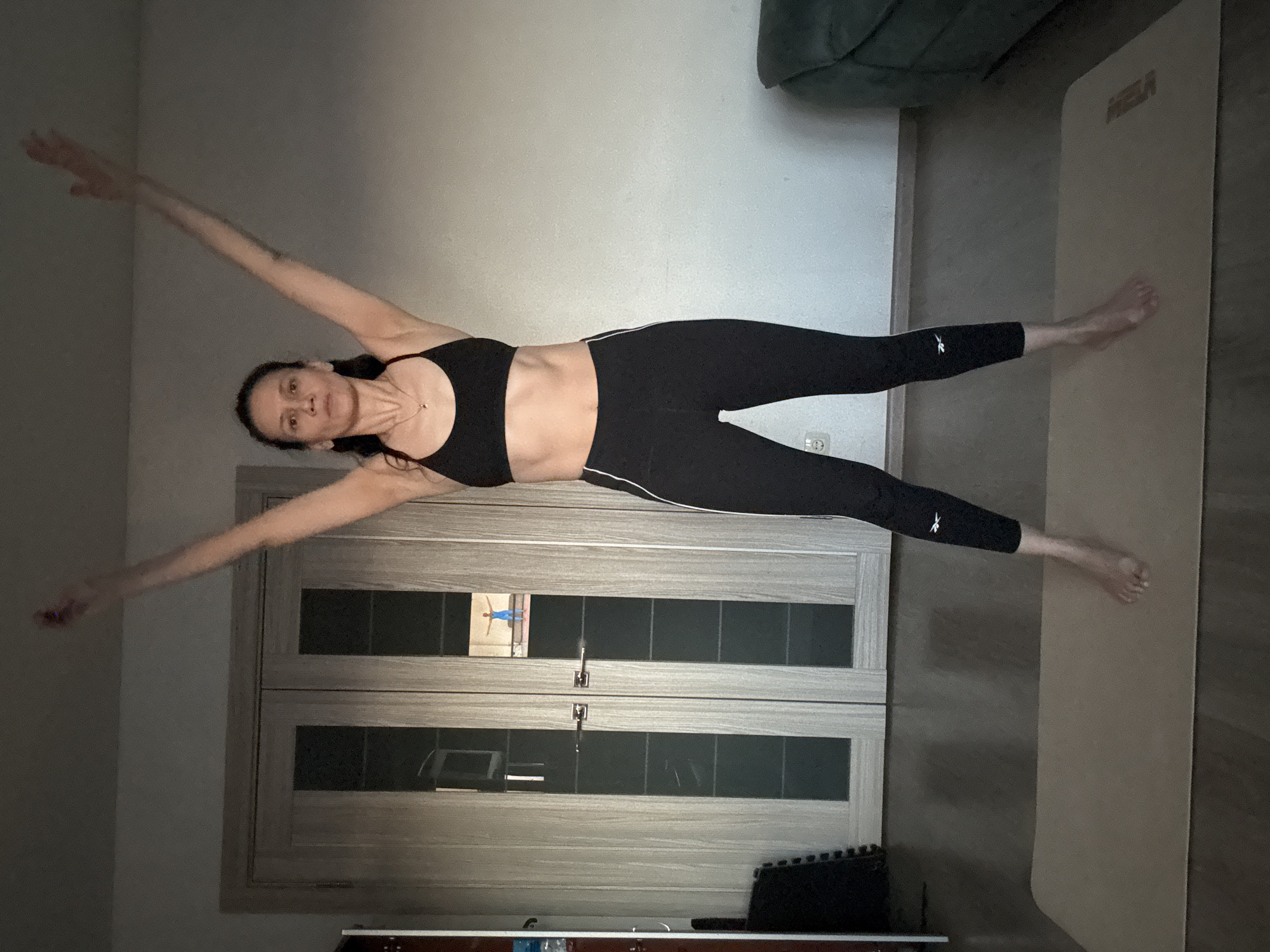 The image captures a woman performing an exercise or yoga pose indoors in Chaikovsky, Russia. The primary subject is a woman with dark hair, wearing a black sports bra and black leggings with white side stripes, extending her limbs horizontally across the frame. She appears focused with a neutral expression.
The setting is a room, likely a home interior, characterized by light-colored walls and a light-toned wooden two-panel door with dark glass inserts. Reflections are visible in the door's glass, including what seems to be a small picture or screen depicting a person in a similar stretched pose. On the floor, a light-colored yoga or exercise mat is present, with the text "MIRA" (possibly a brand name) visible on it. A dark green sofa or armchair is partially visible in the upper right. Other objects include a white electrical outlet on the wall and a dark, ridged object (possibly an exercise accessory) on the floor near the door. The woman's black leggings feature a white Reebok logo on the left calf. Her arms are extended away from her body, and her legs are stretched out. The overall lighting suggests an indoor setting, possibly with artificial light.