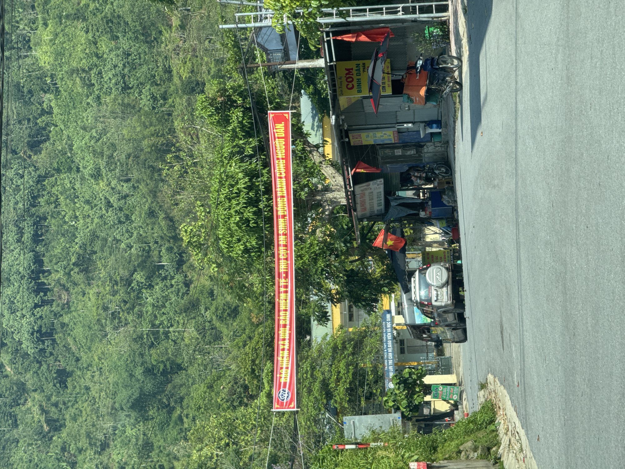 This media file depicts a rural or semi-rural roadside scene in Thông Nguyên Commune, Vietnam, under bright daylight. The image is taken from a slightly elevated perspective, looking down a road that curves slightly to the right in the foreground.
On the left side of the frame, lush, dense green mountains or hills dominate the background, suggesting a mountainous region. Overhead, a long, red banner is strung across what appears to be the road, extending from left to right.
The right side of the image features the asphalt road itself, with a shoulder leading to several single-story buildings or shops. These establishments have corrugated metal roofs and various signs. Parked near these shops are a few motorcycles, one with orange delivery boxes, and further down the road, a silver SUV with a spare tire on its rear is visible. Trees and other vegetation are interspersed among the buildings and along the roadside.
Visible text on the red banner reads: "BẢO HIỂM XÃ HỘI, BẢO HIỂM Y TẾ - TRỤ CỘT AN SINH, ĐỒNG HÀNH CÙNG NGƯỜI DÂN." which translates to "Social insurance, health insurance - Pillars of security, accompanying the people." Another prominent sign, yellow in color, advertises "Quán Hoàng My CƠM BÌNH DÂN PHẦN" (Hoàng My Restaurant Common Rice Portion) and "CƠM XE TAI" (Trucker's Rice) with a phone number "DT: 0948 508 192". Below the main banner, a sign clearly states "TRƯỜNG THCS & THPT THÔNG NGUYÊN" (Thông Nguyên Middle & High School) and "KHU LƯU TRÚ CÁN BỘ, GIÁO VIÊN VÀ HỌC SINH" (Accommodation for staff, teachers, and students), directly confirming the provided location context. Other smaller signs feature text like "BÚN" (vermicelli) and "DA", along with numbers like "5456" on the SUV's license plate.
The scene is tranquil, without any specific activity taking place, conveying a sense of everyday life in a Vietnamese commune.