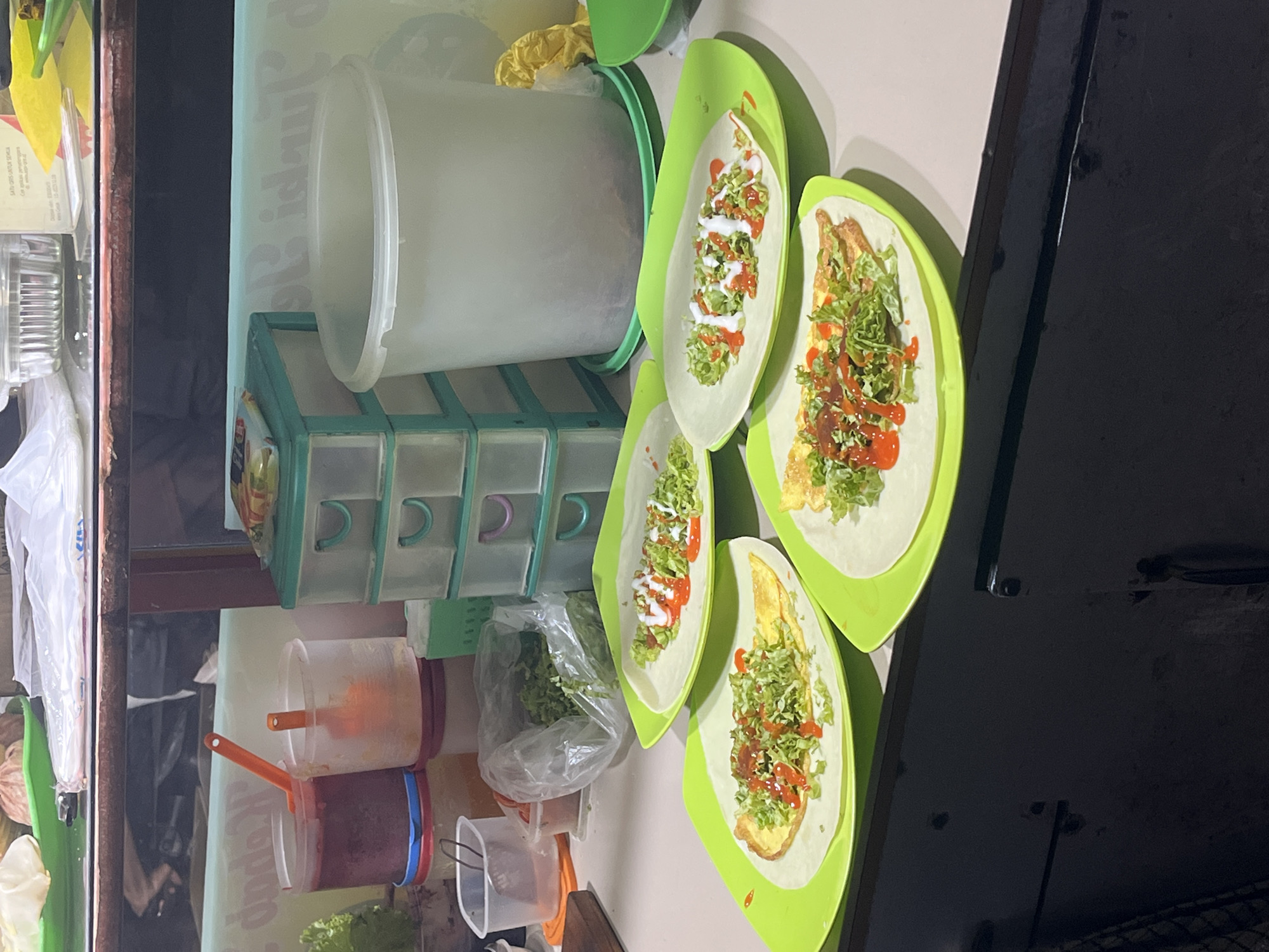 In a street food setting in Lhokseumawe, Indonesia, several dishes are being prepared and displayed. The primary subjects are several wraps, likely burritos or similar, presented on bright green oval plates. Each wrap is filled with shredded lettuce, what appears to be scrambled eggs or omelet pieces, and drizzled with a white sauce (possibly mayonnaise or a sour cream-based sauce) and a red sauce (likely ketchup or chili sauce). Some wraps also have small red pieces, possibly tomatoes or chili.
To the left of the wraps, there is a light-colored plastic container with a green lid, possibly for ingredients. Below this, a teal-colored organizer with multiple small drawers is visible, containing various items. Further left, there are several clear plastic containers, some filled with orange and red sauces, each with a small orange-handled spoon or ladle. A plastic bag filled with shredded green vegetables, likely lettuce, is also present.
The scene suggests a food stall or small eatery, likely operating during the day or early evening, as there is artificial lighting illuminating the area. The background is a light-colored wall with some faint text that is not clearly legible. The overall impression is one of food preparation and service in a casual, outdoor or semi-outdoor environment. The focus is on the fresh ingredients and the finished wraps, indicating a meal being offered to customers.