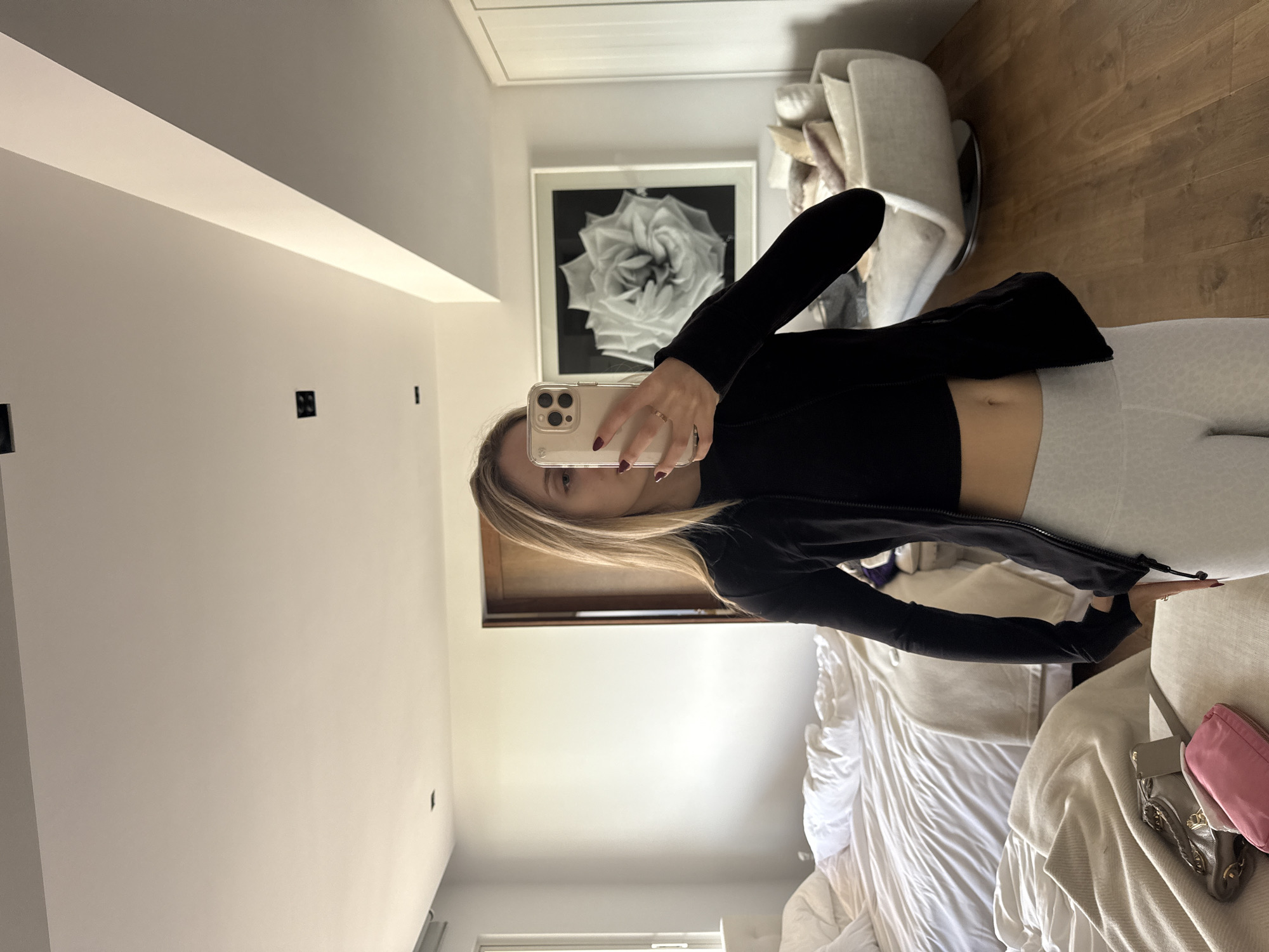 A blonde female, visible from the chest down, is centered in a modern, well-lit interior room located in Mexico City, Mexico. She wears a black long-sleeved athletic jacket over a black crop top, paired with light-colored, patterned athletic leggings. Her left hand, adorned with dark red nail polish and a ring, is visible. She holds a light-colored smartphone with her right hand, obscuring most of her face, and appears to be taking a mirror selfie.
The room features light-colored walls, recessed ceiling lighting, and wooden flooring. To the left, a large, unmade bed with white bedding is visible, alongside a light-colored throw draped over a foreground surface. On this surface, a grey textured bag with gold hardware and a pink pouch are present. A window with a white frame is situated above the bed. To the right, a large framed black and white abstract floral artwork hangs on the wall, with a modern, light-colored upholstered lounge chair positioned beneath it. White paneled cabinetry or a closet is visible further to the right. A dark wooden door frame is centrally located in the mid-background.