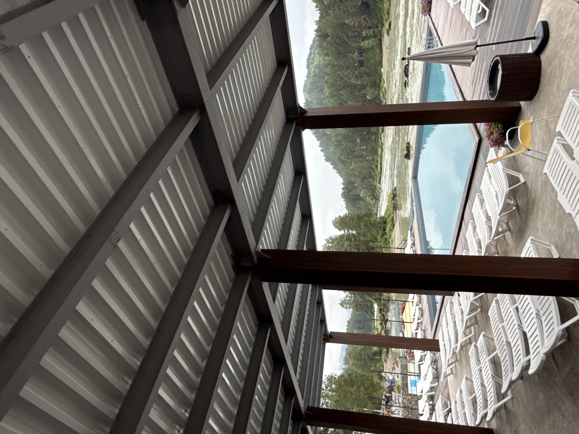 The image depicts an outdoor swimming pool area at a resort, likely located in or near Barangol, Russia, given the context of a river and mountainous terrain. The scene is viewed from under a covered patio with a corrugated metal roof supported by dark brown wooden posts and dark gray metal beams. Numerous white plastic lounge chairs are arranged in rows on a paved surface, some facing a large rectangular swimming pool filled with blue water. To the right of the pool, a closed beige umbrella stands next to a dark wooden hot tub. Beyond the pool, a wide, muddy river flows through a forested valley with steep, tree-covered hills in the background. The sky is overcast, suggesting a cloudy day. In the distance, on the riverbank to the left, a few people are visible near some yellow boats, indicating ongoing activity. The overall atmosphere is one of a resort leisure area.