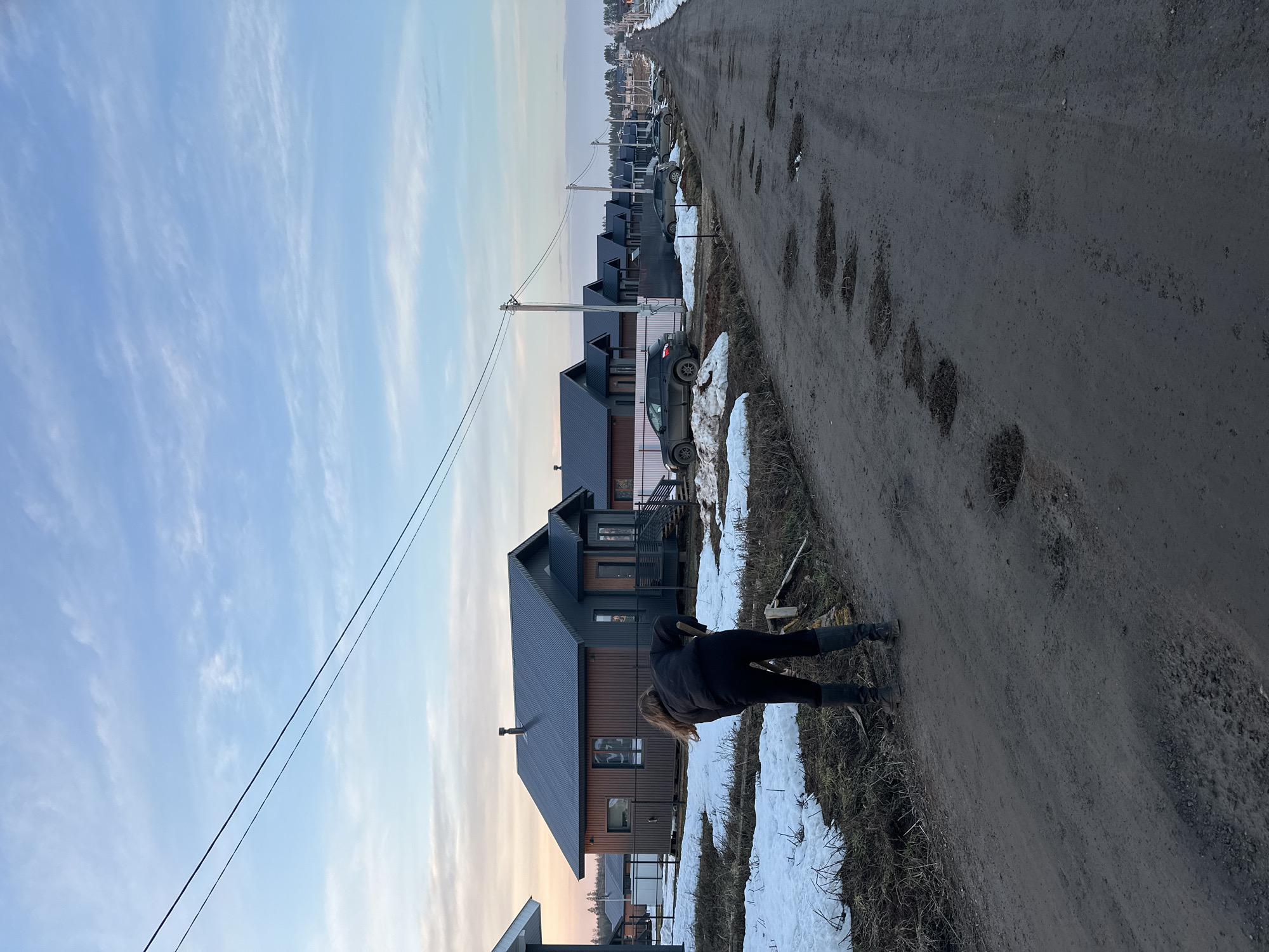 The image depicts a street scene in Подшивалово, Russia. The street is unpaved and riddled with potholes. A person is bent over near the edge of the road. The person is wearing dark clothing and boots. The person has shoulder-length blond hair. Adjacent to the road are modern-style houses. Snow patches remain along the side of the road and around the houses. A car is parked near one of the houses. Power lines run along the street. The sky is light blue with some clouds.