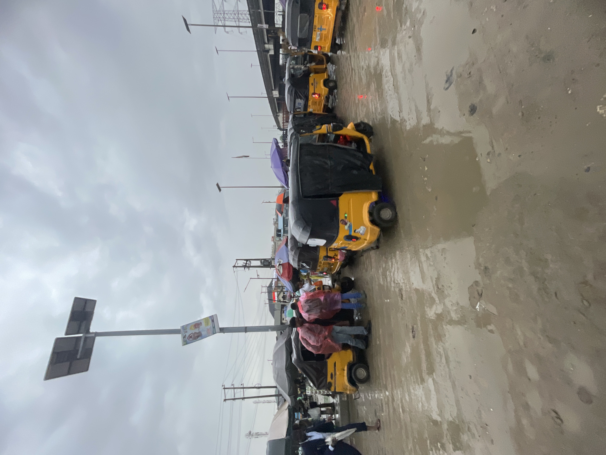 The image shows a street scene in Lekki, Nigeria, on a rainy day. The ground is wet and reflective. Several yellow and black auto-rickshaws ("keke napeps") are lined up along the road. People are seen near the vehicles, some holding umbrellas. Overhead power lines and a signpost are visible. An elevated highway overpass is in the background. The sky is overcast with gray clouds.