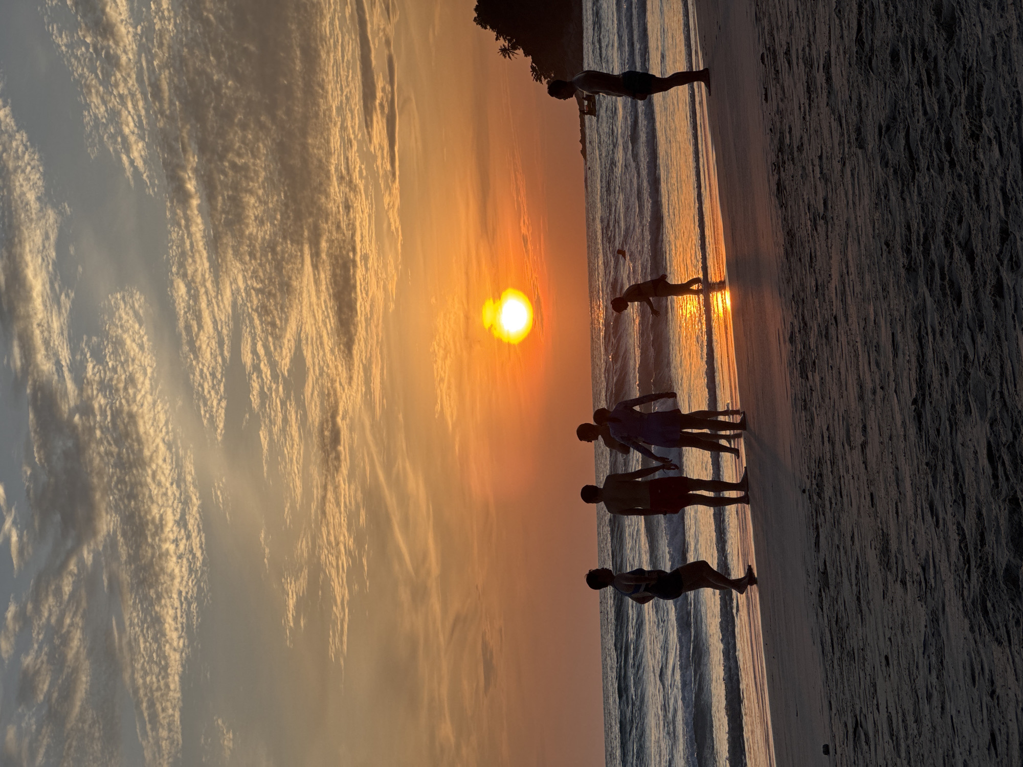 A group of people are silhouetted on a beach in Karon, Thailand, during sunset. The sun, a bright orange orb, is low on the horizon, casting a warm glow across the sky and water. Wispy clouds are scattered across the sky, catching the last rays of sunlight. The waves of the Andaman Sea gently lap against the shore. The sandy beach is dark and textured, with footprints visible.
In the foreground, a person stands with their back to the camera, facing the sea. To their left, a group of three people stand close together, one in a blue swimsuit and two in swimwear. Further down the beach, a child walks towards the water. In the distance, a person stands in the water, and another is further out to sea.
The overall scene is one of a peaceful and beautiful sunset on a popular Thai beach.