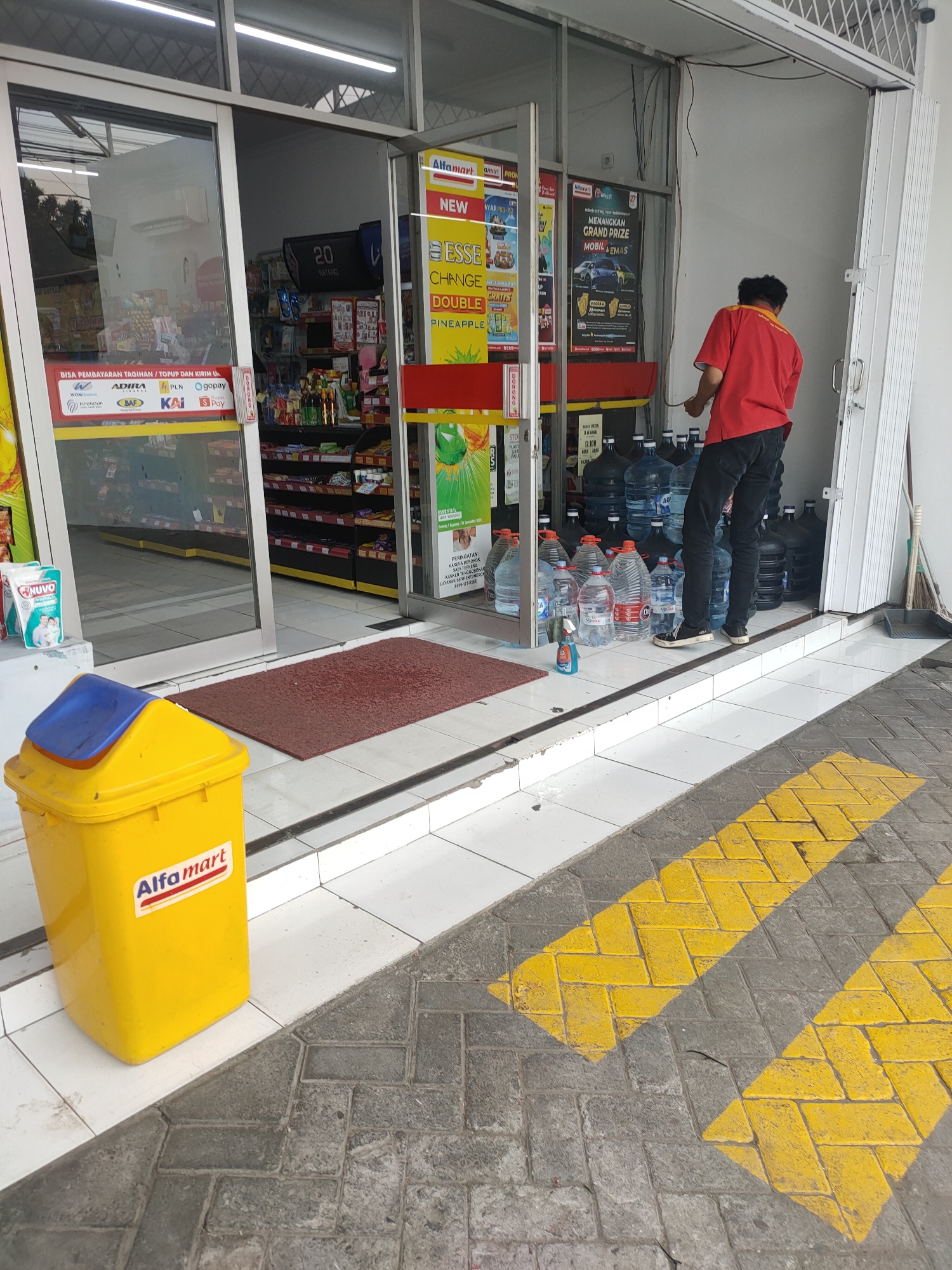 A man in a red shirt and black pants stands in the doorway of an Alfamart store in Kanigoro, Indonesia. He is facing the interior of the store, with his back to the camera, and appears to be adjusting something near a stack of bottled water. Inside the store, shelves are stocked with various products, and a digital display shows "20 DATANG". Outside the store, to the left of the entrance, is a yellow trash can with a blue lid, bearing the Alfamart logo. A red doormat is placed on the white tiled floor in front of the entrance. The ground outside the store is paved with interlocking gray bricks, marked with yellow diagonal lines.