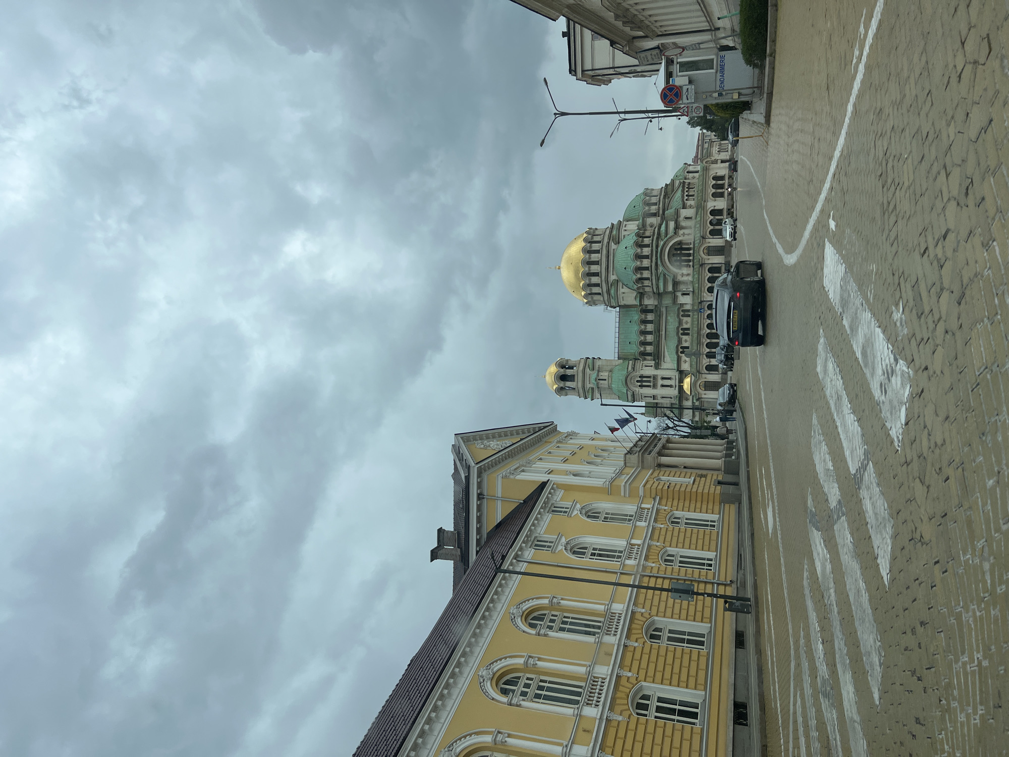 The image captures a street view in Sofia, Bulgaria, featuring the Alexander Nevsky Cathedral prominently. The cathedral's distinctive green domes, topped with gold, stand out against a cloudy sky. A yellow building with arched windows is visible on the left. Cars are present on the street, and a crosswalk is in the foreground. The street is paved with yellow brick. Other buildings are visible along the street. A street lamp is present.