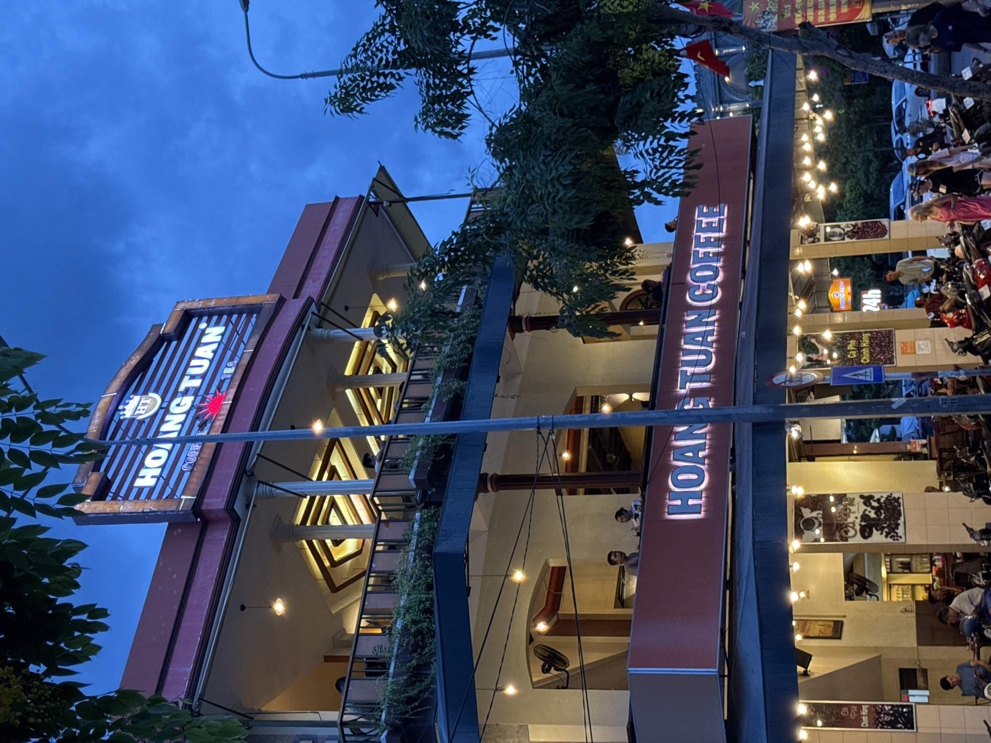 In Nha Trang, Vietnam, at dusk, a multi-story building with signage reading "HOANG TUAN COFFEE" is illuminated by string lights and the building's internal lighting. The establishment appears to be a coffee shop with outdoor seating on multiple levels, evidenced by people visible on the balconies. Below the coffee shop, a street scene is depicted with numerous parked motorcycles and a few people walking. The sky is a deep blue, indicating the transition from day to night.