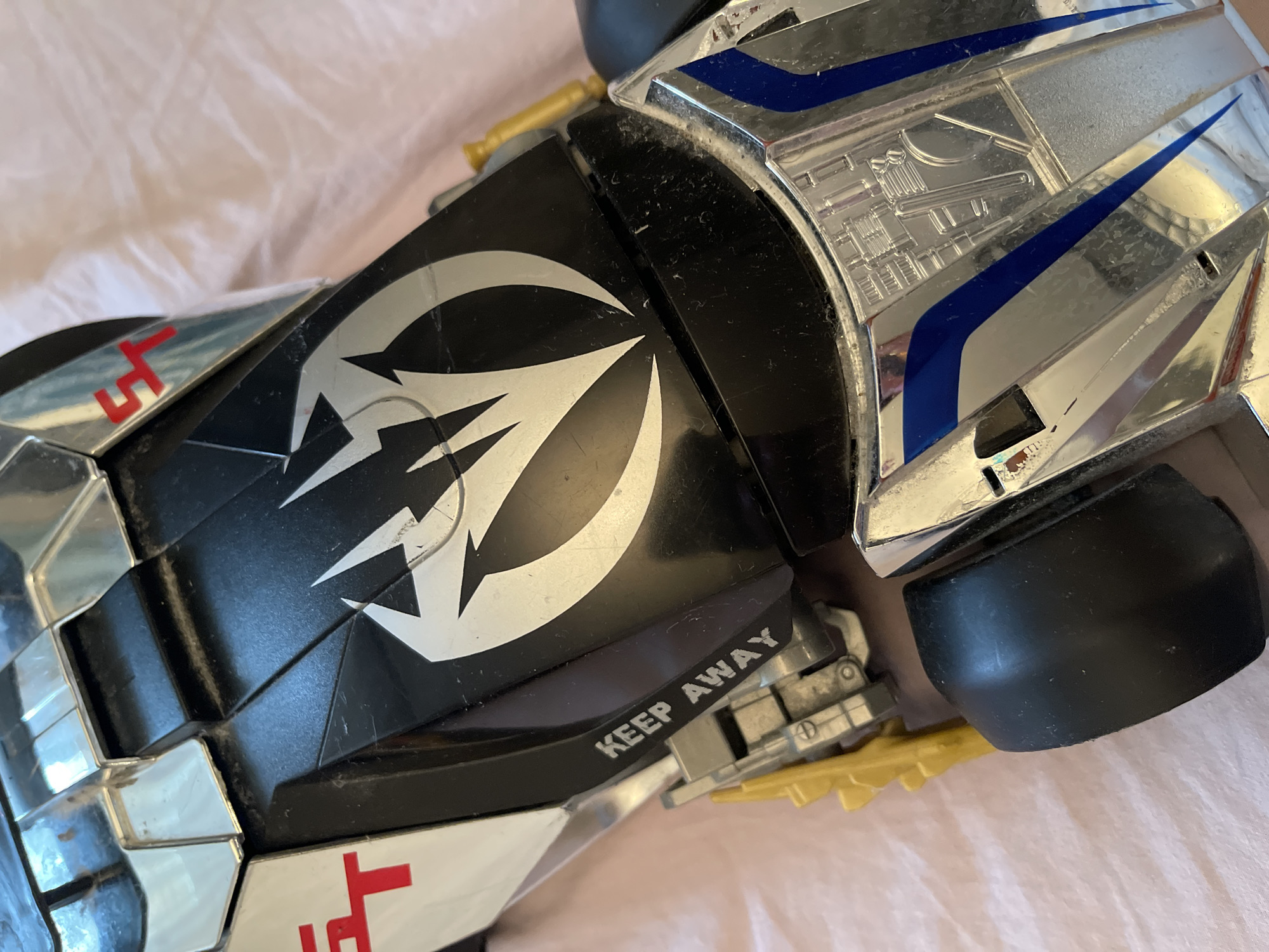 The image shows a close-up of a toy vehicle on a pinkish surface. The vehicle is primarily black and silver with blue accents. A silver logo with claw-like shapes is prominently displayed on the black portion. Text "KEEP AWAY" is visible on the side of the vehicle. Red markings are also present. The vehicle has black tires. There is no evidence to suggest that the location is Moscow, Russia.