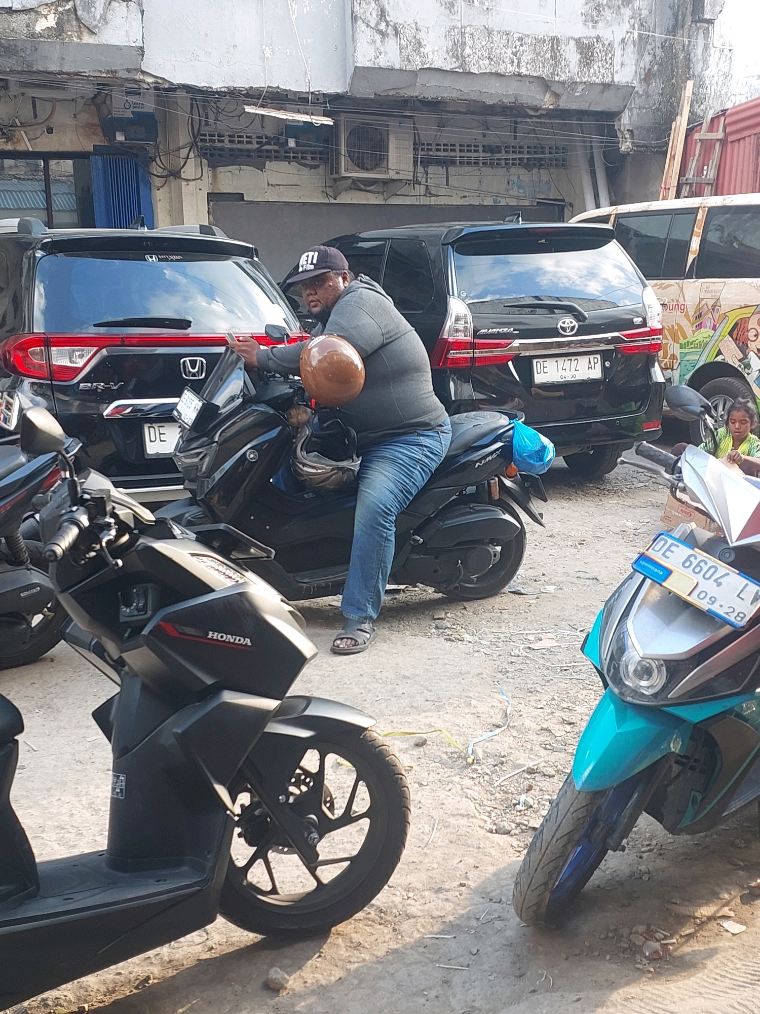 A man wearing a black cap and a grey hoodie is sitting on a black scooter. He is holding a brown helmet and looking at his phone. He is wearing blue jeans and sandals. There are several cars and other scooters parked around him. In the background, a young girl is sitting on the ground. The scene appears to be in a city street with buildings and a weathered facade. The license plate of the scooter is partially visible, showing "DE 6604 LY". The environment is outdoors, on a paved surface, with some debris on the ground. The presence of multiple vehicles suggests a parking area or a busy street.