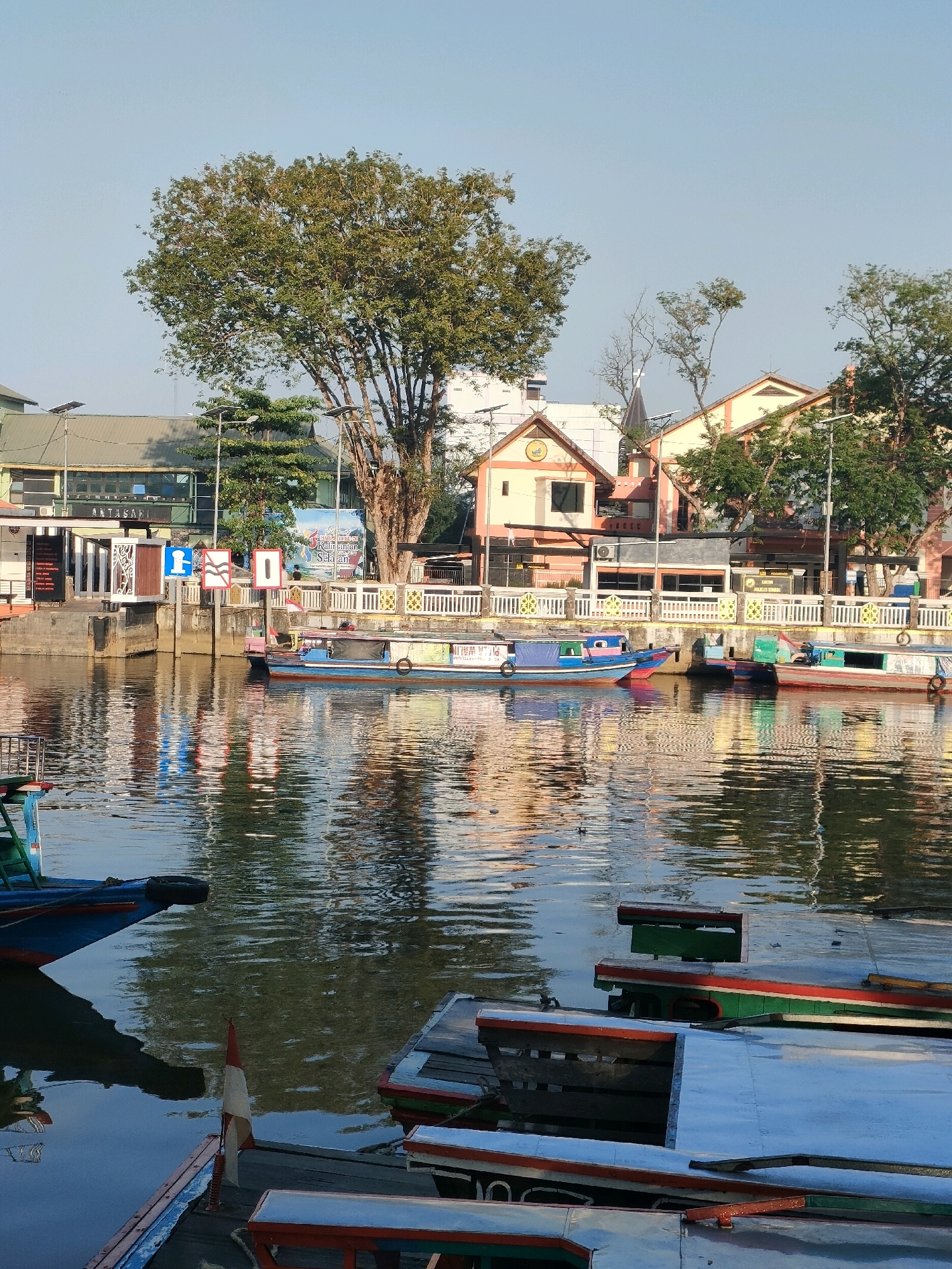 The image depicts a scene along the Martapura River in Banjarmasin, South Kalimantan, Indonesia, likely during daytime given the clear sky and bright lighting. The foreground features several moored boats, some with wooden benches and red trim, docked along a waterfront. The water's surface is rippled, reflecting the surrounding buildings and trees.
In the mid-ground, a narrow canal or river is lined with a pedestrian walkway and a railing. Several passenger boats, painted in various colors including blue, red, and green, are docked along this waterway. One boat is particularly prominent, with blue and red accents and a covered seating area.
The background reveals a collection of buildings, characteristic of a city environment. These structures have tiled roofs and appear to be a mix of commercial establishments and possibly residential buildings. Signage is visible on some of the buildings, including one that reads "ANTASARI". Trees, including a large, leafy specimen dominating the left side of the frame, are interspersed among the buildings, casting shadows on the water. The overall impression is of a busy riverside urban area.