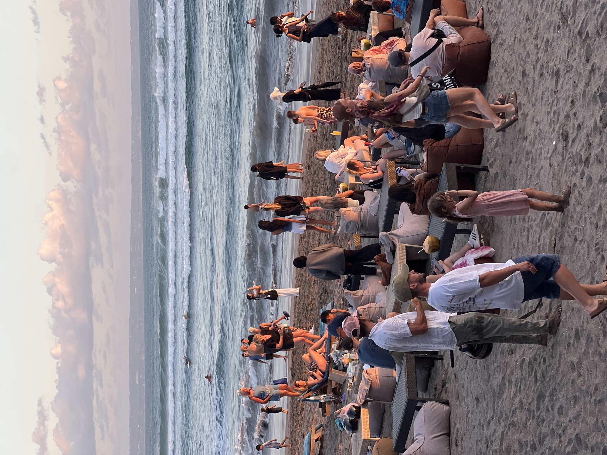 The image depicts a beach scene in North Kuta, Indonesia, with numerous people engaged in various activities. The beach is sandy, with several low tables and beanbag chairs set up, suggesting a dining or lounging area. In the background, the ocean has moderate waves, and several individuals are in the water, some with surfboards. The sky is partly cloudy, with soft pink and orange hues, indicating it is likely sunset or sunrise.
In the foreground, a group of people are seated or standing around the tables. A man in a white shirt and olive pants stands near a table. Another man in a white t-shirt with a graphic print and dark shorts is walking past. A young girl in a pink dress stands on the sand.
Further back, a variety of individuals are seen interacting and relaxing. Some are seated on beanbags, some are standing and talking, and others are walking along the beach. A woman in a blue denim outfit is standing, and a woman in a black dress is holding a white purse. People are also visible in the middle ground, some near the water's edge. The overall atmosphere appears to be relaxed and social.