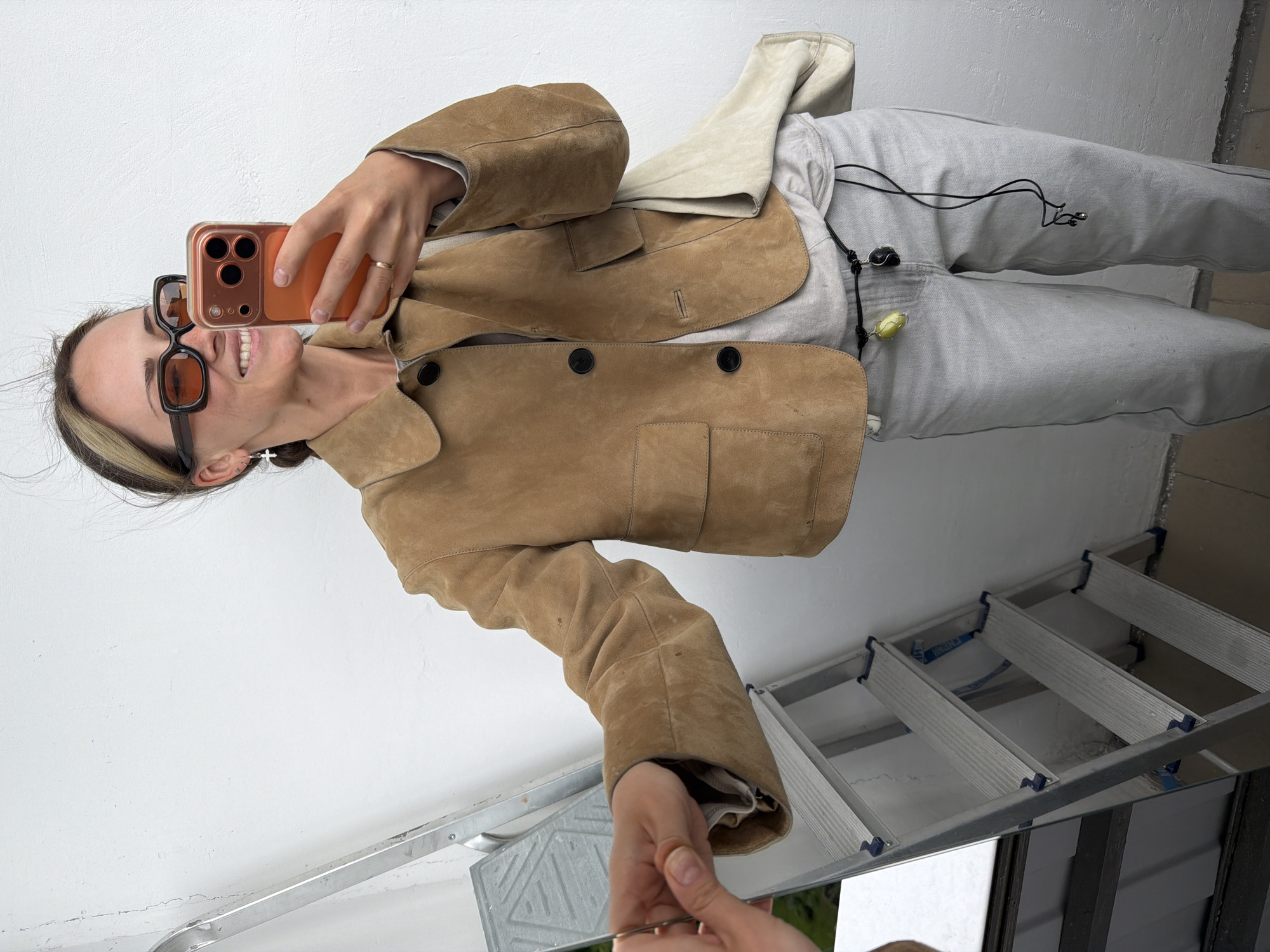 A woman stands on a folding aluminum ladder inside a building, possibly in the city of Khimki, Russia. She is holding a mirror in her left hand and a cell phone in her right hand, taking a selfie. She is wearing sunglasses, a light brown jacket, and light grey pants. Her hair is pulled back, with a light streak in the front. The environment around her consists of a white wall and the ladder.