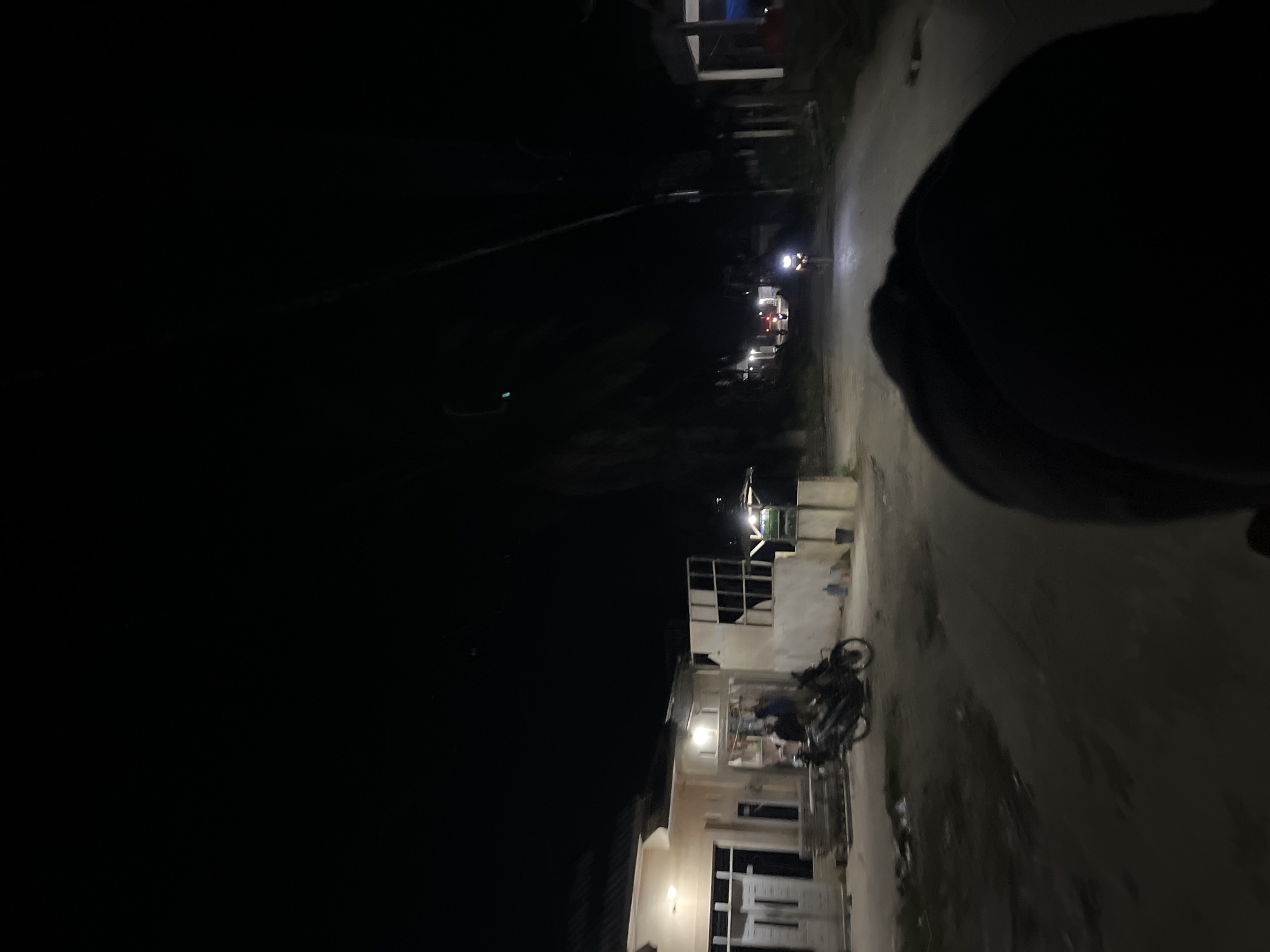 The image depicts a street scene at night in Landuh, Indonesia. The perspective suggests the photo was taken from a moving vehicle, likely a motorcycle, as a dark shape, possibly a helmet or shoulder, occupies the foreground on the right. The street is paved and illuminated by the headlights of a motorcycle in the distance and lights from buildings.
Several buildings are visible on the left side of the street, some appearing to be under construction or in a state of disrepair. One building shows a white facade with open window frames and visible interior structures. A motorcycle is parked in front of this building. Further down the street, more lights indicate other structures and possibly more activity.
On the right side of the street, the road surface is visible, reflecting the ambient light. The overall impression is a quiet, residential or semi-commercial area at night, with minimal pedestrian activity. The time of day is clearly nighttime due to the darkness and artificial lighting. There are no visible signs of text in the image. The atmosphere is subdued, typical of a nocturnal urban or semi-urban environment.