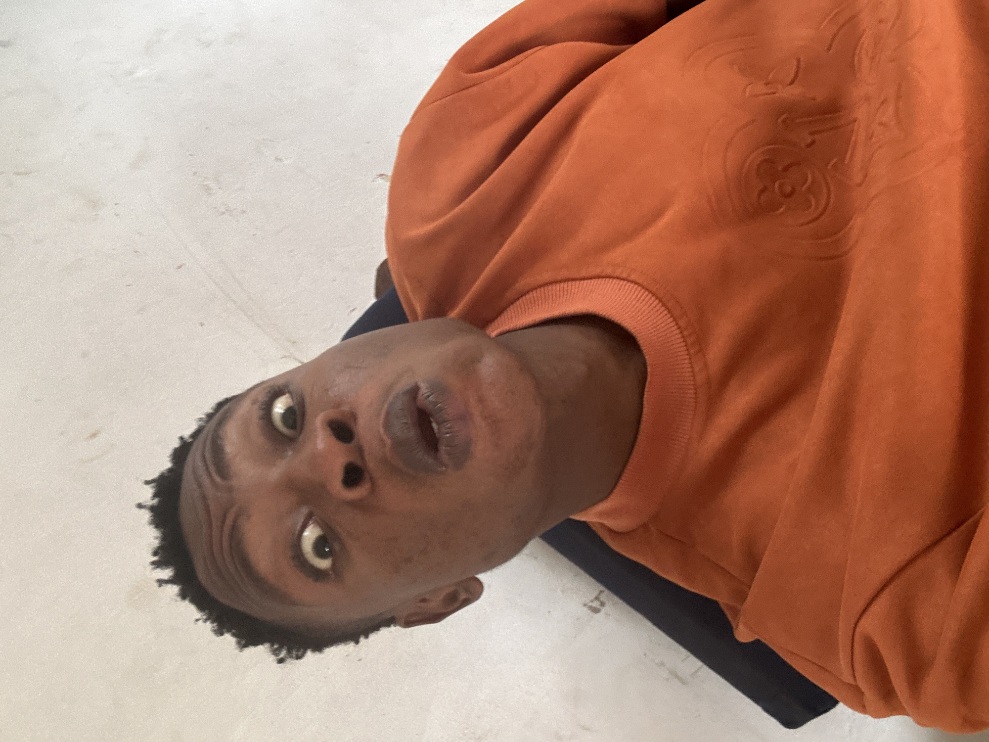 An eye-level medium shot captures a Black male from the chest up, looking directly into the camera with a surprised or shocked expression. His eyes are wide open, and his mouth is slightly agape. He has short, dark, textured hair and appears to be in his late teens or early twenties. He is wearing an orange crewneck sweatshirt with a tonal embossed design on the chest, possibly a logo or pattern. The background is a textured, off-white wall with some faint smudges and marks. The lighting is bright, casting soft shadows. The man is seated, and a dark object, possibly the back of a chair or a dark garment, is visible behind his left shoulder. The image seems to be taken indoors.