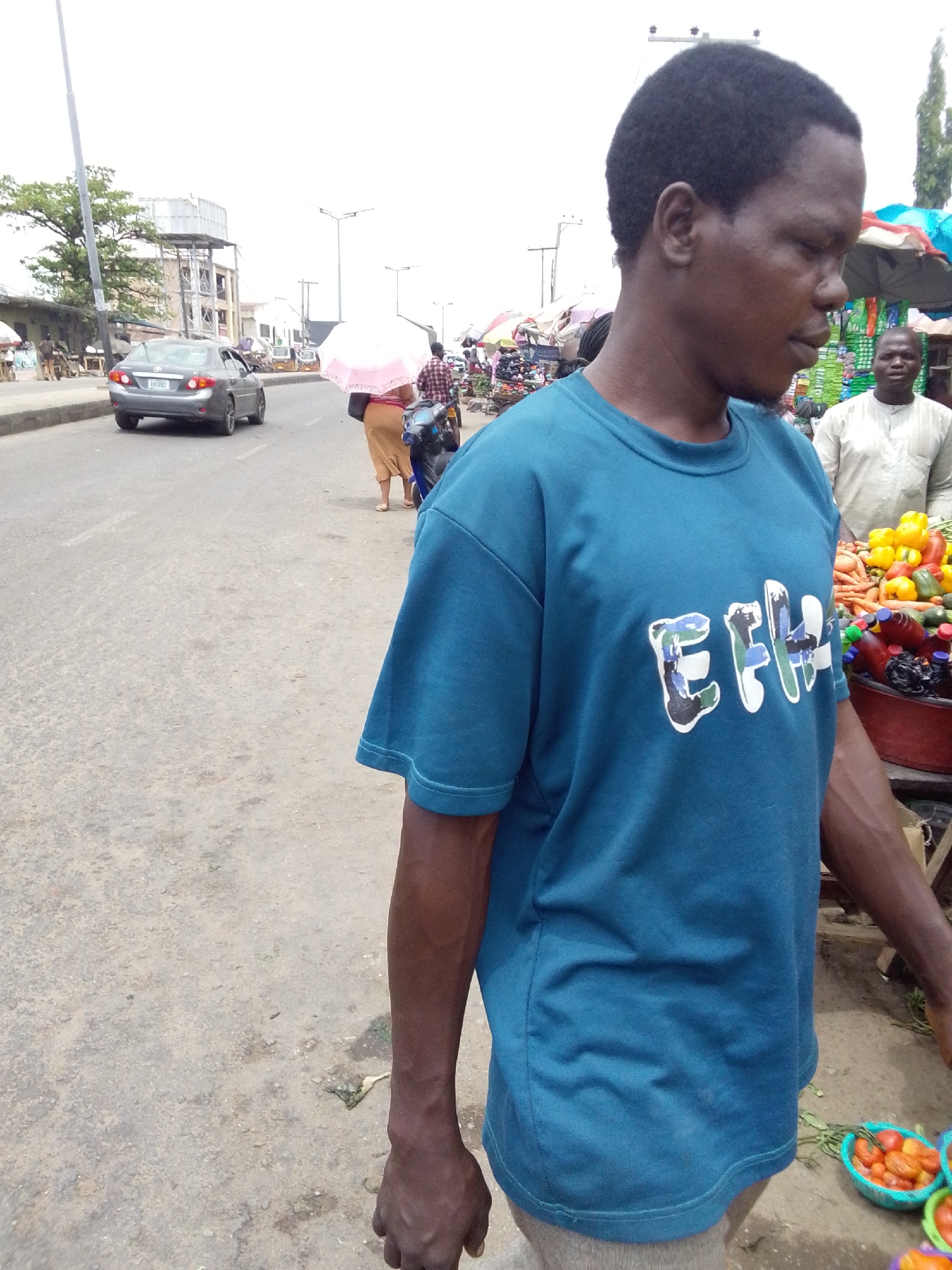 This image depicts a bustling street market scene, likely in Lokoja, Nigeria. The primary subject is a man of African descent, positioned in the right foreground and looking downwards to his right. He wears a plain teal or dark blue short-sleeved t-shirt with a stylized, camouflage-patterned text "EFH" (or possibly "EHH") across the chest. He has short, dark, curly hair and dark skin.
In the background to the right, market stalls are visible, overflowing with colorful fresh produce such as bright yellow bell peppers, orange carrots, and red tomatoes contained in plastic bowls. Another man, wearing a light-colored traditional garment, stands behind one of these stalls.
To the left of the frame, a paved road extends into the distance. A silver sedan car is seen driving away from the viewer. Further down the road, a person walks holding a light pink umbrella. The street is lined with various buildings and utility poles, including a prominent structure with an elevated water tank on top of a building.
The scene is set during the daytime under an overcast sky, suggested by the diffused lighting and the absence of harsh shadows. The ground consists of a mixture of dirt and worn pavement. The overall impression is that of a lively urban or semi-urban marketplace demonstrating daily commercial and street activities.