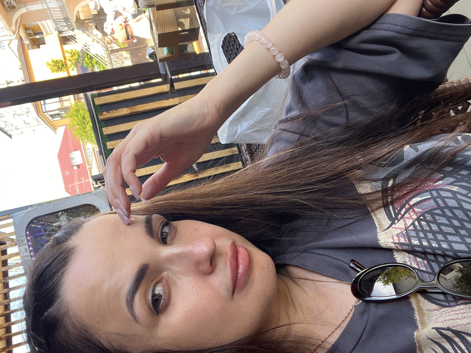 A woman with long brown hair is seated outdoors, partially obscured by a wicker chair. She is wearing a dark gray t-shirt and a rose quartz beaded bracelet on her right wrist. Sunglasses hang from a delicate necklace around her neck. Her right hand is raised to her forehead, and her left arm is resting on the armrest of the chair. The background shows a building with orange and yellow facades, a red-tiled roof, and trees. Some people are visible in the background, appearing to be walking. The lighting suggests it is daytime.