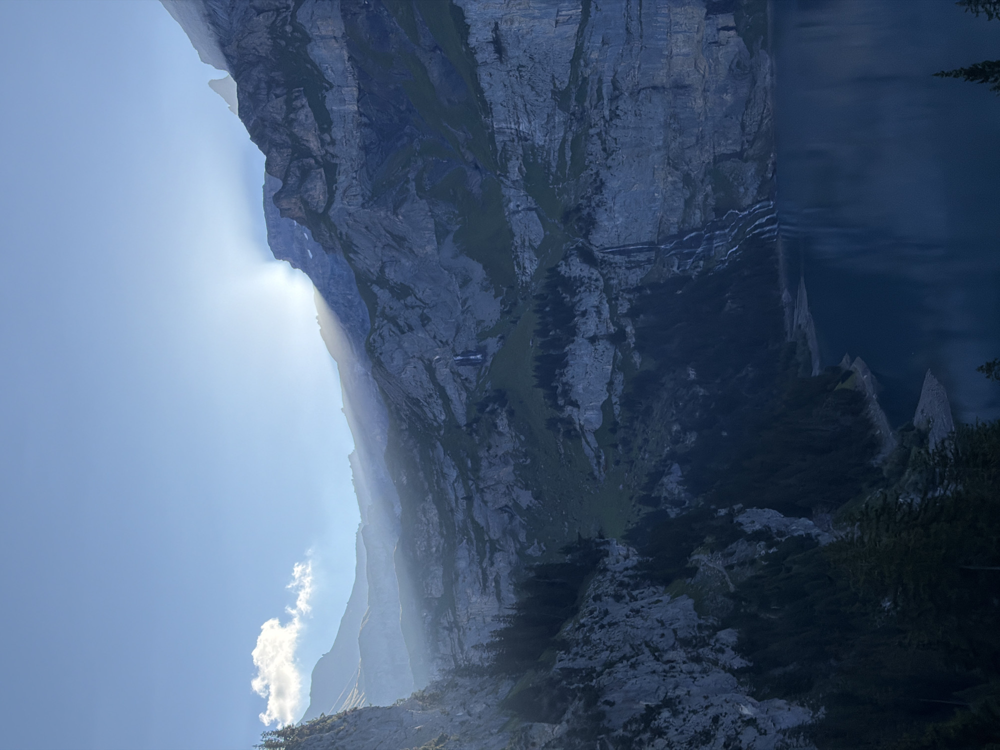The image captures a breathtaking mountain landscape with a deep blue lake in the foreground, reflecting the rugged peaks. The sun is partially obscured by the mountains, casting dramatic rays and creating a misty haze across the scene. Waterfalls cascade down the sheer rock faces, adding to the dramatic natural beauty. Pine trees dot the steep slopes leading down to the lake's edge. The overall atmosphere is one of serene grandeur, indicative of a high-altitude environment in the Swiss Alps, likely Oeschinensee, near Kandersteg, Switzerland.