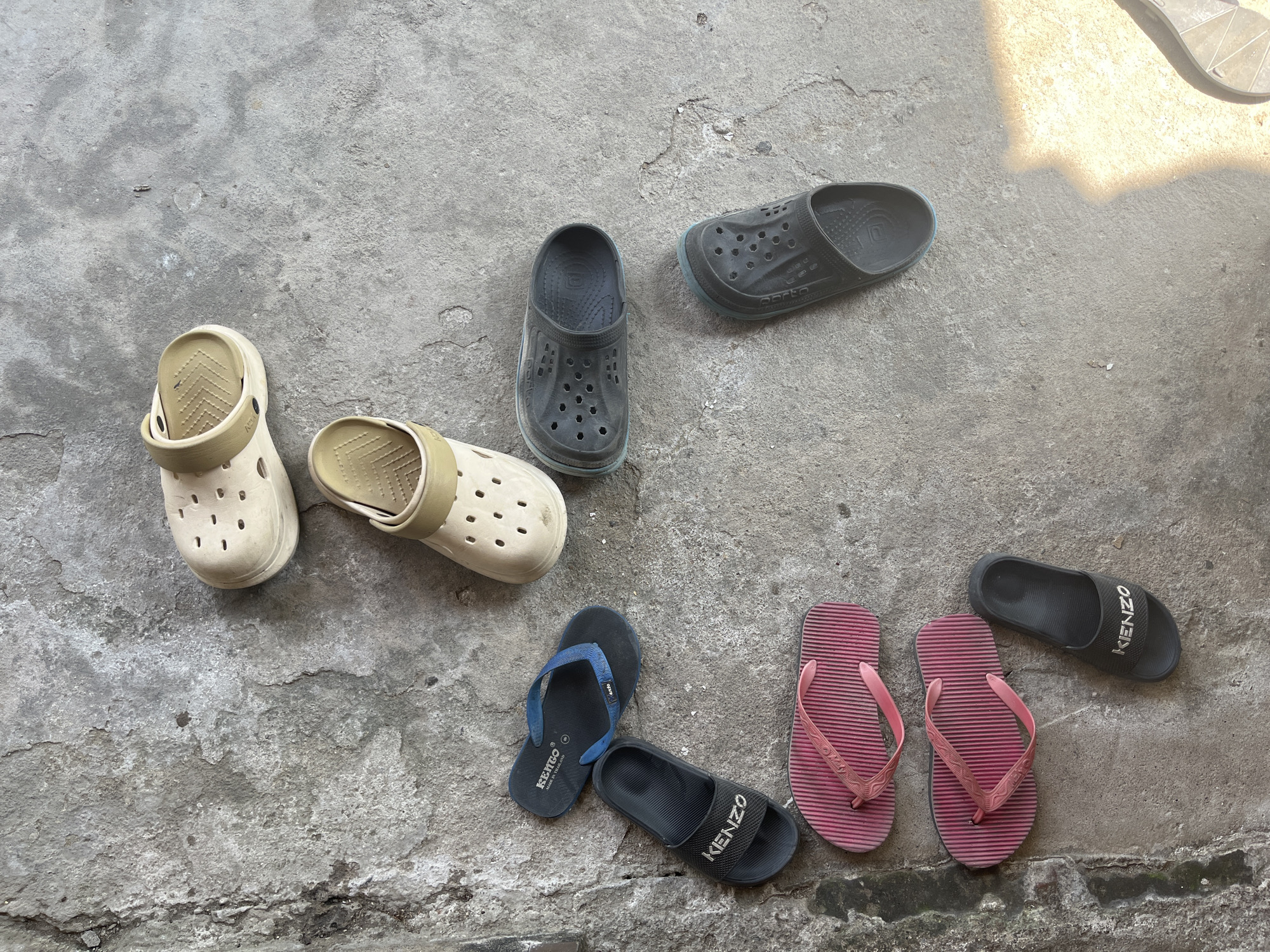 This is a top-down, forensic photograph of several pairs of footwear scattered on a rough, concrete surface. The scene appears to be outdoors, possibly in an alleyway or a yard, with patches of shadow suggesting the presence of an overhead structure or the sun. The concrete is cracked and weathered, with some debris visible.
The footwear includes:
- Two pairs of light beige clogs with ventilation holes.
- Two pairs of dark gray clogs with ventilation holes.
- One pair of blue and black flip-flops with the brand "KENZO" visible on the strap.
- One pair of black slides with the brand "KENZO" prominently displayed on the strap.
- Two pairs of pink flip-flops with textured soles and straps.
The arrangement of the footwear is haphazard, with no clear pattern. There are no people or animals present in the image, and no actions or interactions are depicted. The focus is solely on the collection of shoes and the surrounding environment. The location is described as Bumi Mulyo, Indonesia, though this information is provided as context rather than directly observable in the image.