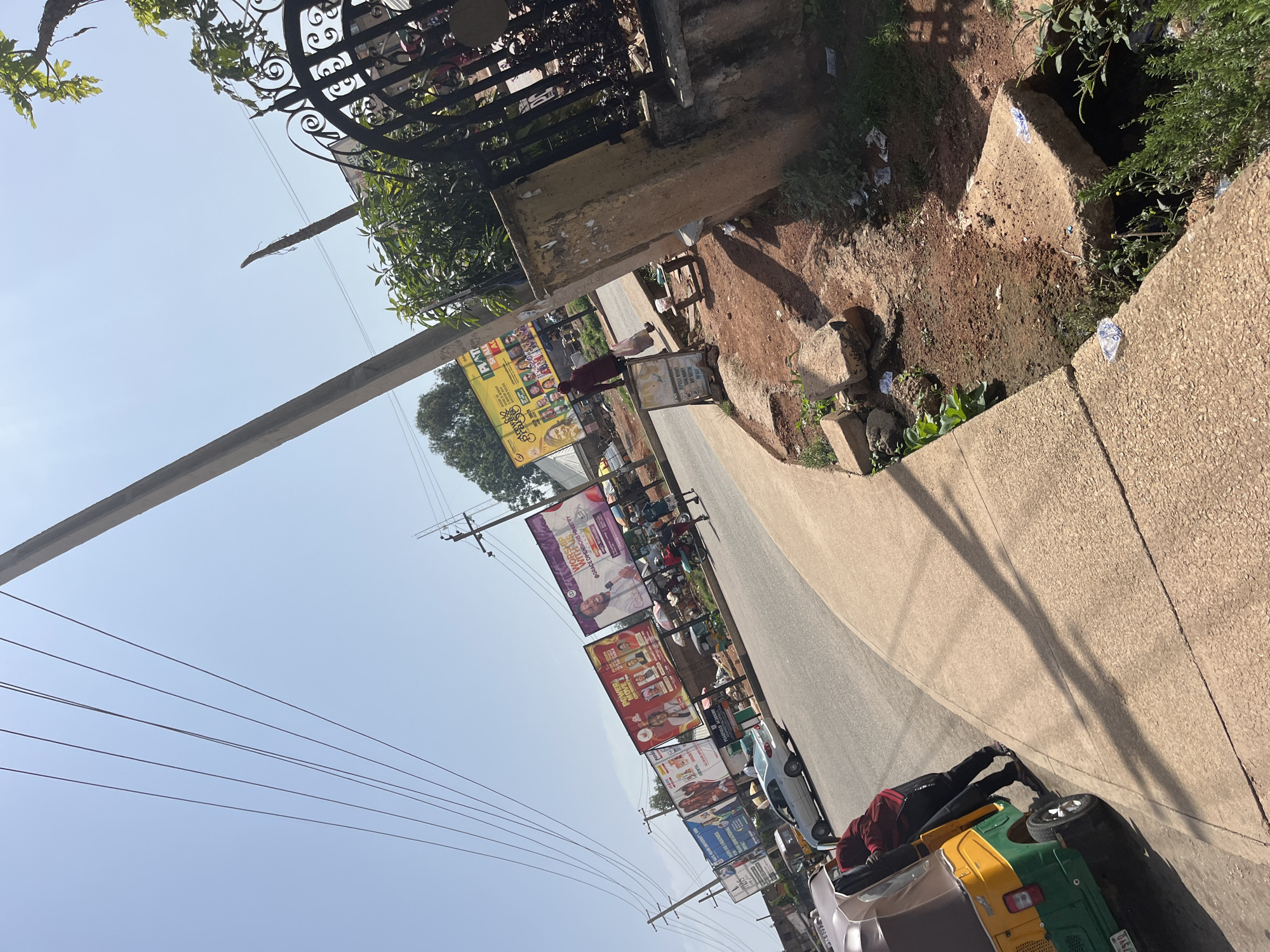 The image depicts a street scene in Gura Top, Nigeria, under a clear, bright sky. A paved road curves into the distance, lined with billboards displaying various advertisements. On the left side of the road, a yellow and green auto-rickshaw is parked, with a person in a dark red jacket and black pants leaning against its open door. A silver sedan is also visible on the road. The right side of the road features a raised concrete sidewalk bordering a ditch filled with debris and vegetation. Beyond the ditch, a black wrought-iron gate is partially visible, with a concrete structure and lush green foliage next to it. Several utility poles with overhead wires are scattered throughout the scene, and trees with dense green leaves are visible in the background. The ground appears to be a mixture of soil and scattered litter.