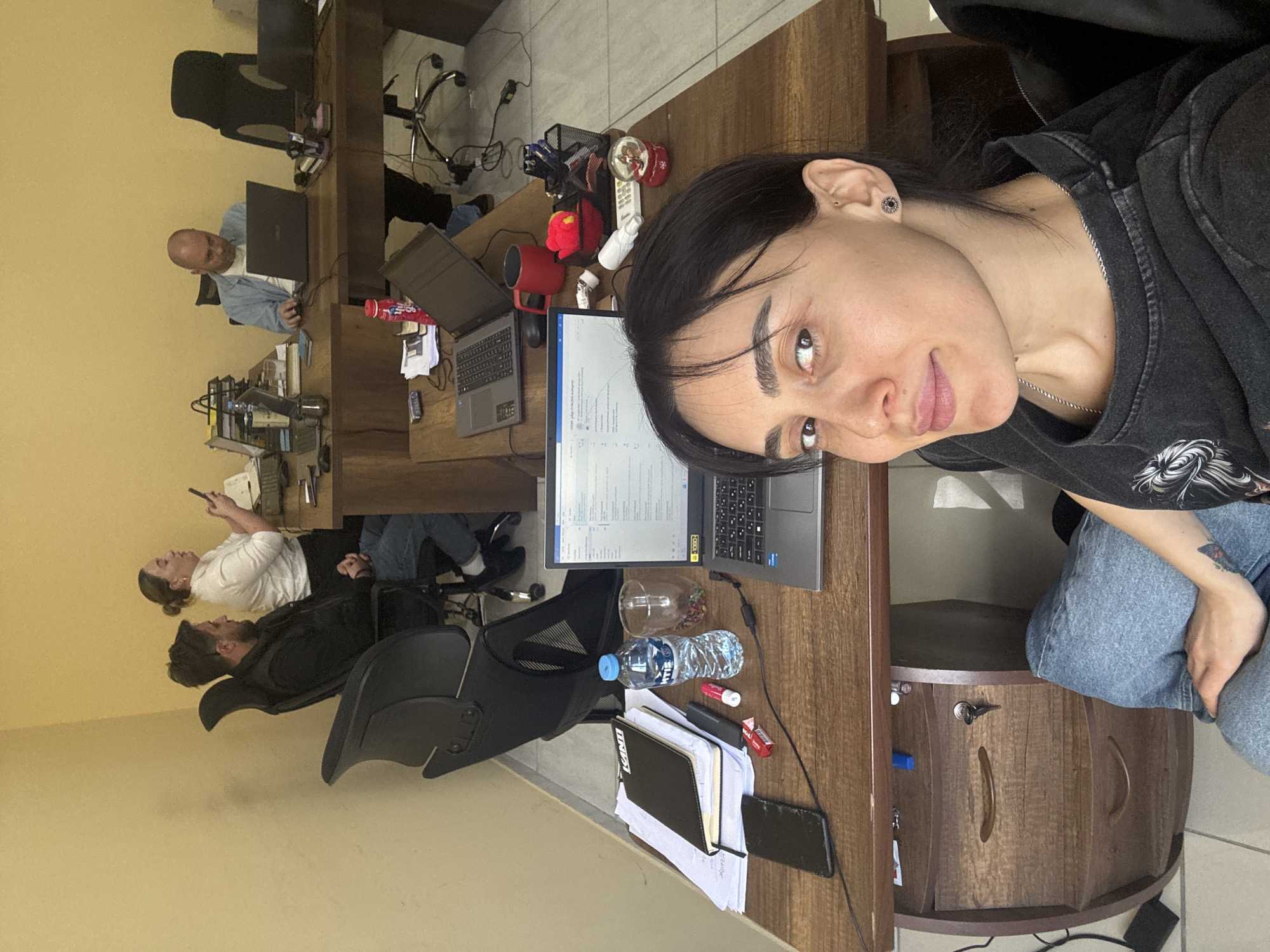An office scene in Tbilisi, Georgia, displays a group of people working at desks. A woman in a dark hoodie and jeans is seated at the desk closest to the camera and appears to be taking a selfie. On her desk are a laptop, a water bottle, notebooks, and a smartphone. Further back, a bald man in a blue shirt is seated at another desk, also working on a laptop. Beyond them, two other people are visible: a man in a dark jacket and a woman in a white shirt. One is looking at a phone. Desks are wooden. The office has a light-colored wall and tiled floor.