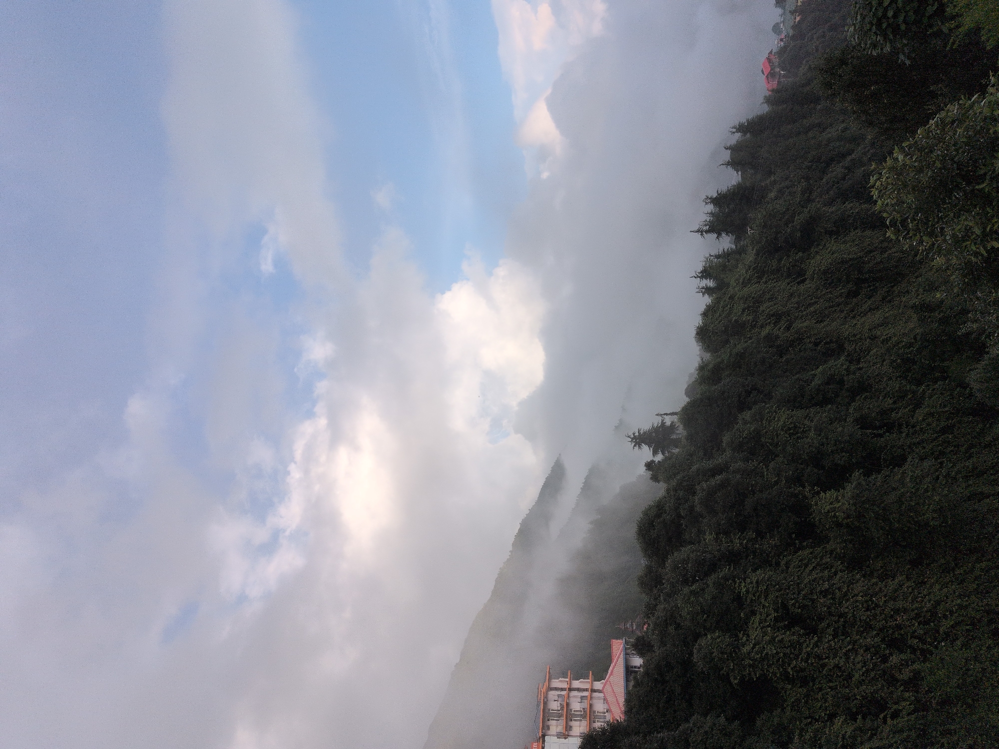This image captures a view of a mountainous landscape in the city of Haini, India. A multi-story building with a red roof and white walls is situated on the left side of the frame, partially obscured by mist and fog. Dense green trees cover the slopes of the mountains, extending across the foreground and middle ground. The sky above is a mix of grey and blue clouds, with patches of sunlight breaking through. The overall scene is serene and slightly atmospheric due to the presence of fog.