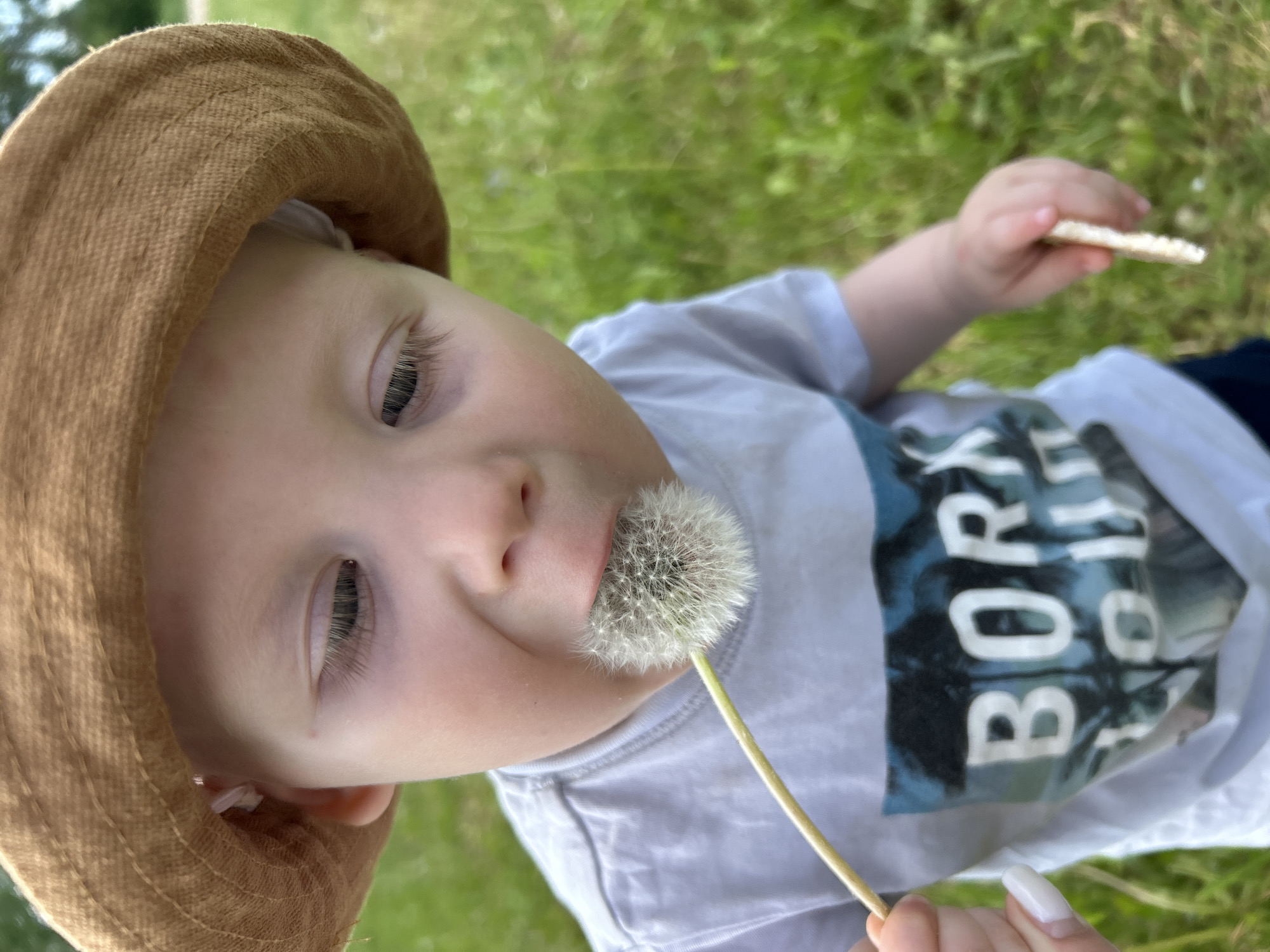A young child wearing a brown hat and a gray t-shirt with a blue and white graphic is holding a dandelion in their mouth. They are also holding a rice cake in their other hand. The child is lying in green grass. The image does not provide any information about the location in the city of Фоки, Russia.