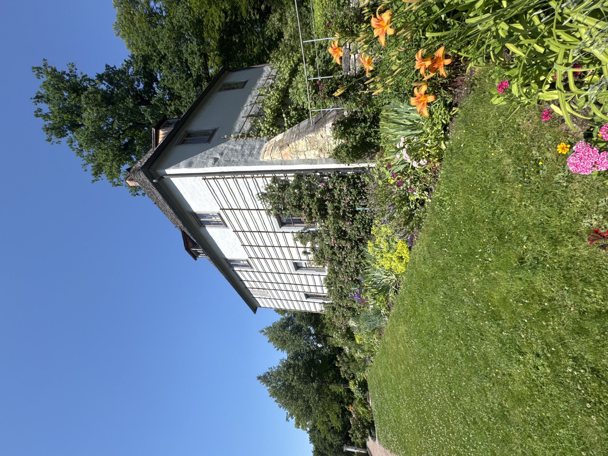 The image shows the Goethe National Museum in Weimar, Germany. The building is a two-story white structure with a gray roof and dormer windows. A trellis-like structure with wooden bars is attached to the side of the building. The building is surrounded by a lush garden with a variety of flowers, including orange daylilies and pink carnations. A green lawn with small white flowers slopes down from the garden. Trees and a clear blue sky are visible in the background.